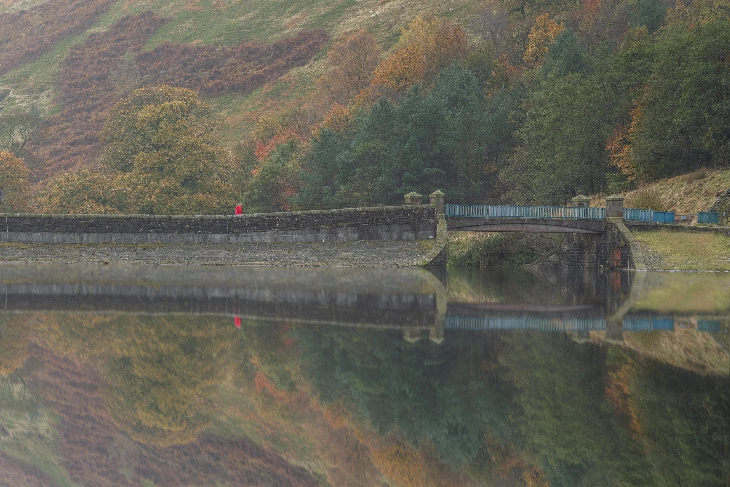 Stone reservoir embankment bridge in Naden Valley reflected in still water with rich autumn trees on the hillside.