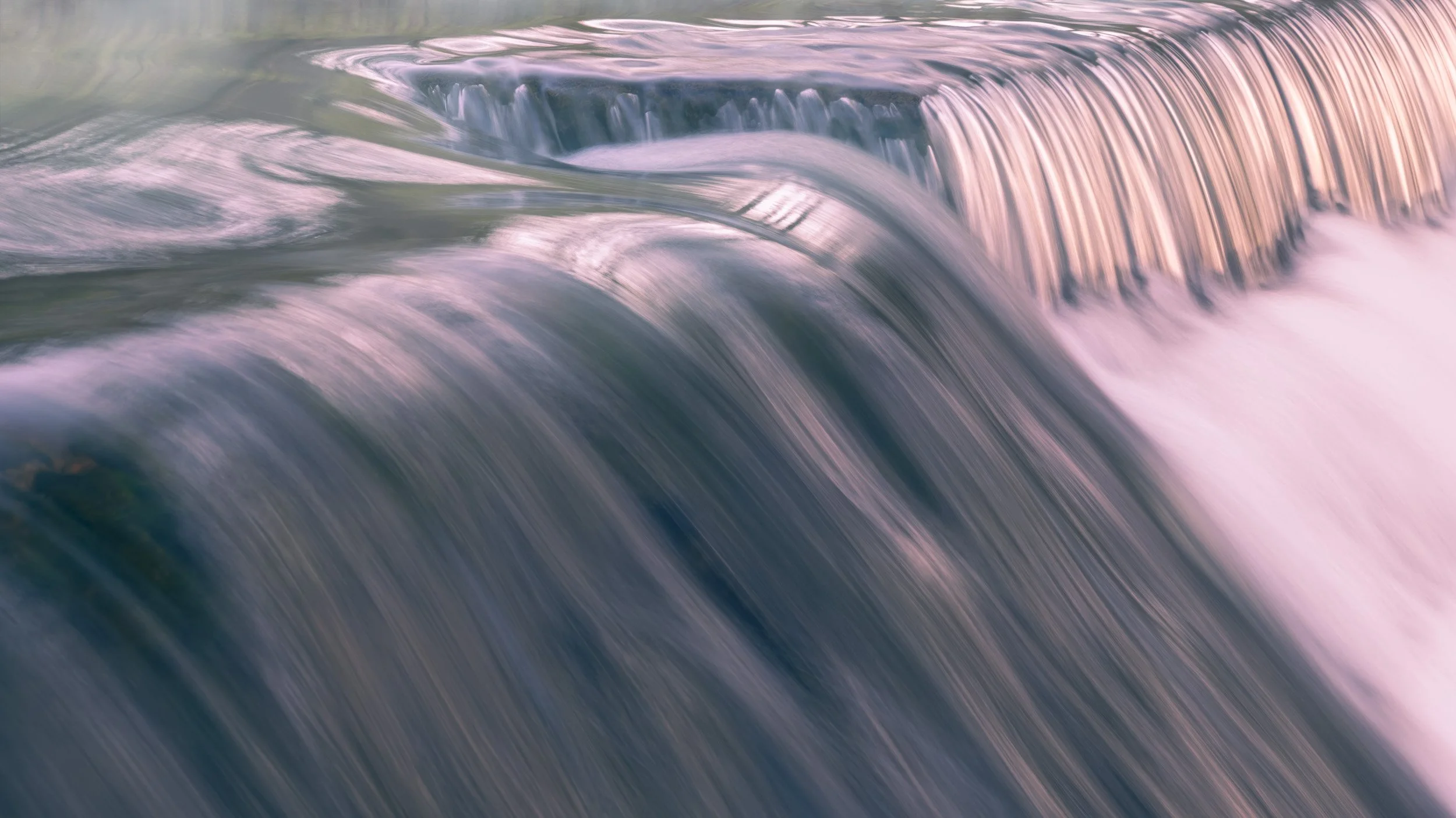 A long-exposure landscape photo capturing smooth, silky water cascading gently over a stone weir on the River Roch with soft violet and golden reflections on the river surface.