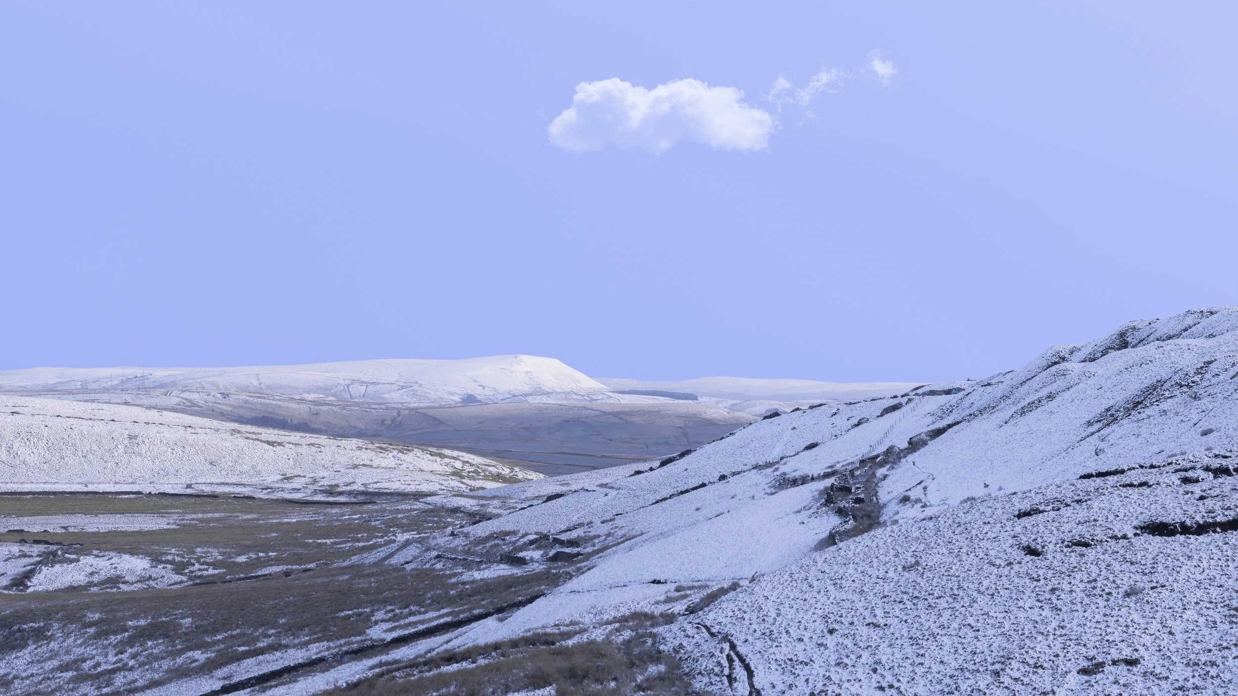 nowy slopes of Foe Edge on Scout Moor near Crag Quarry, looking out toward the snow-capped silhouette of Pendle Hill under a clear blue sky with a single cloud.