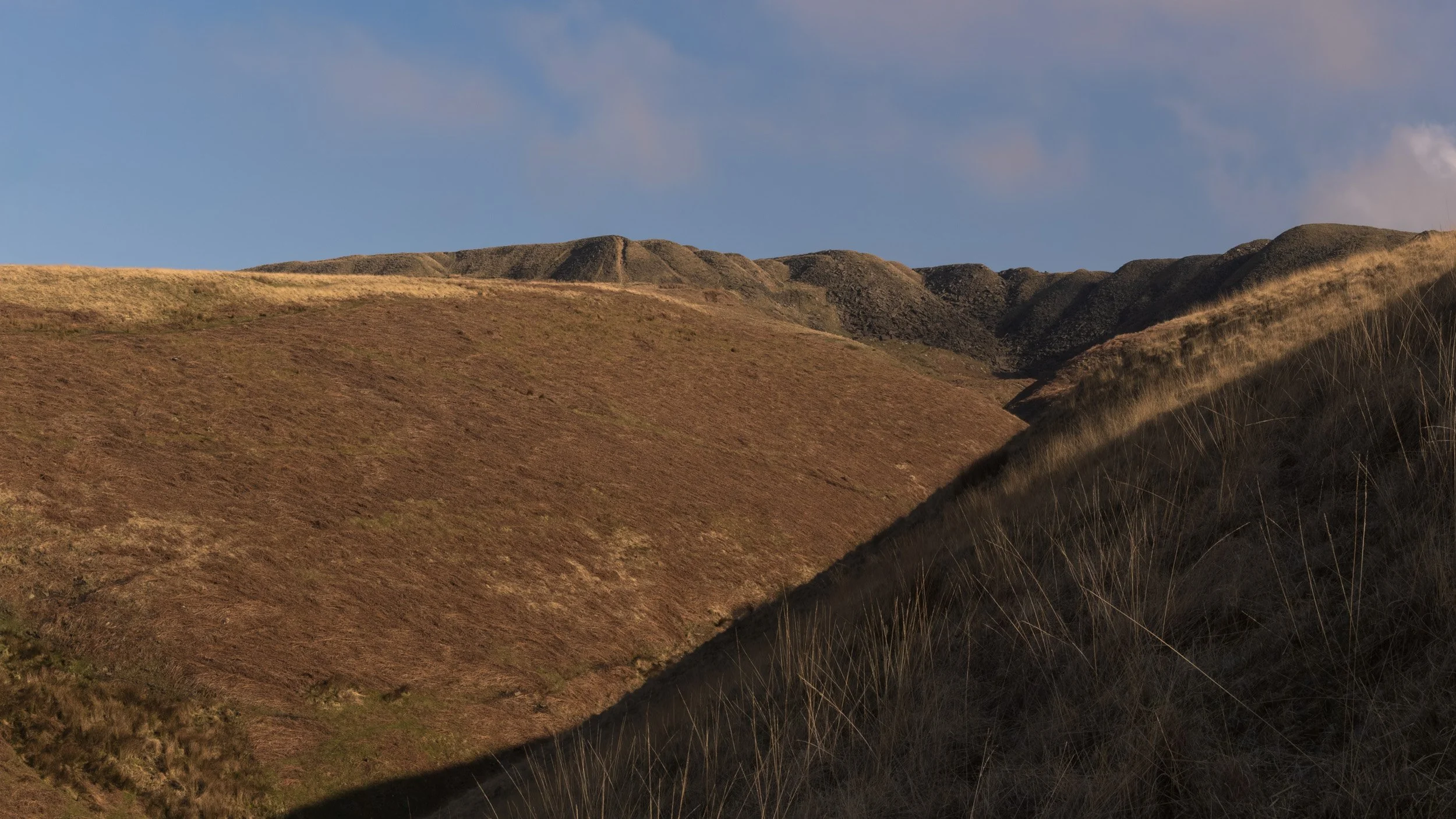 Grassy slopes and deep shadows of Ding Quarry on Rooley Moor near Scout Moor, bathed in late afternoon sunlight against a soft blue sky.