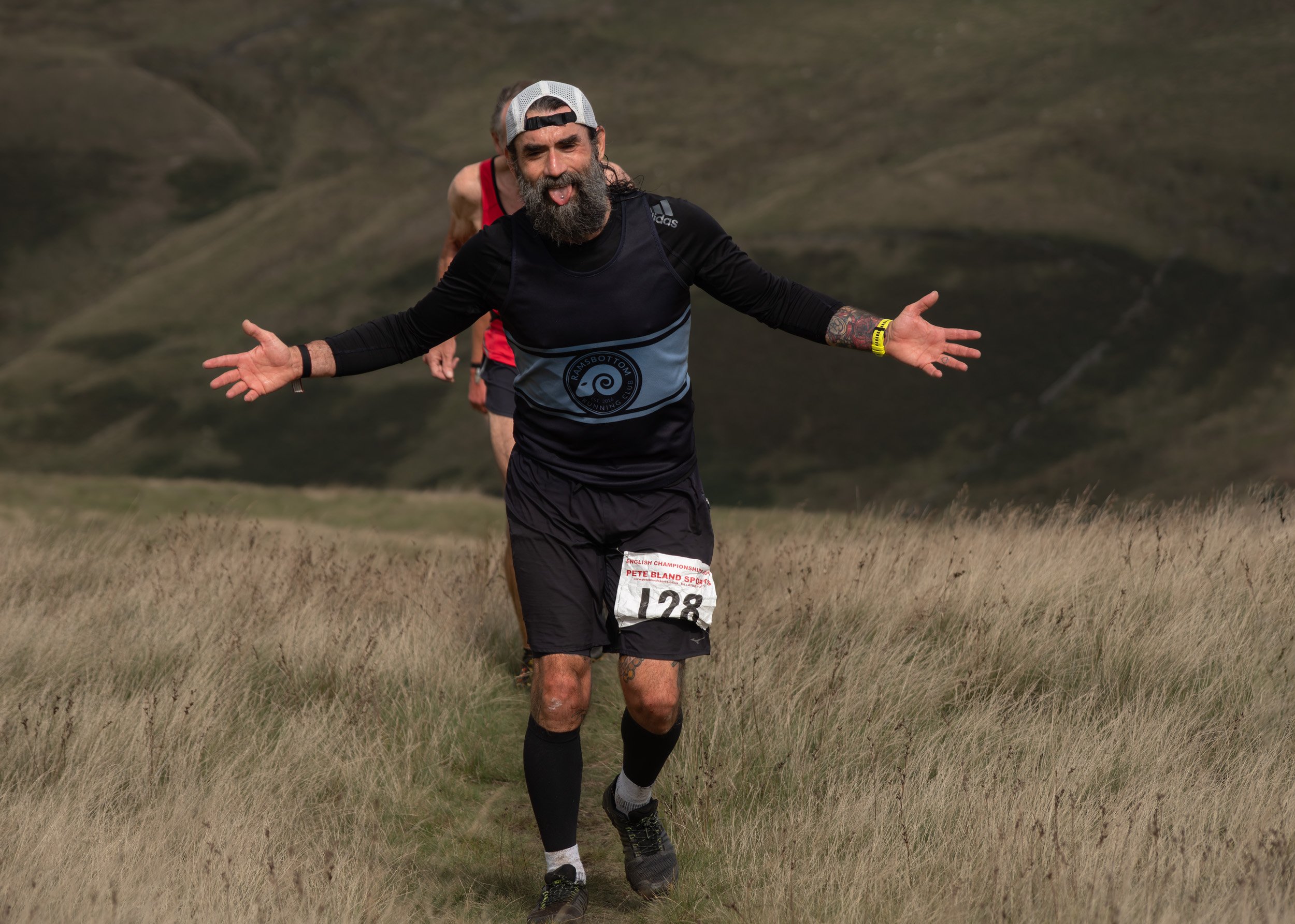 Bearded fell runner wearing race number 128 and a cap running with arms spread wide and tongue out on the Whittle Pike course.