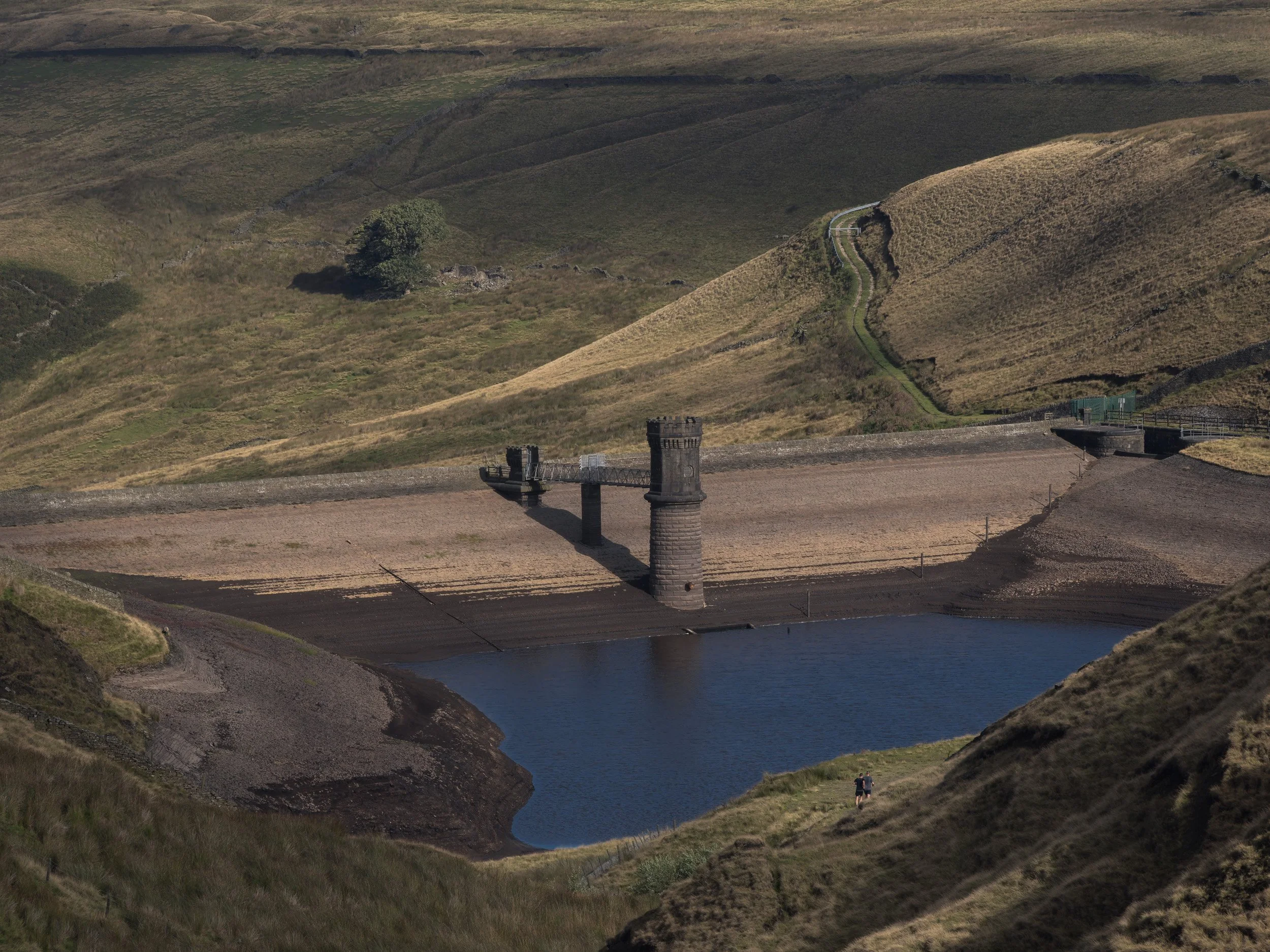 High-angle view of Scout Moor High Level Reservoir at low water level with runners silhouetted on the foreground path.