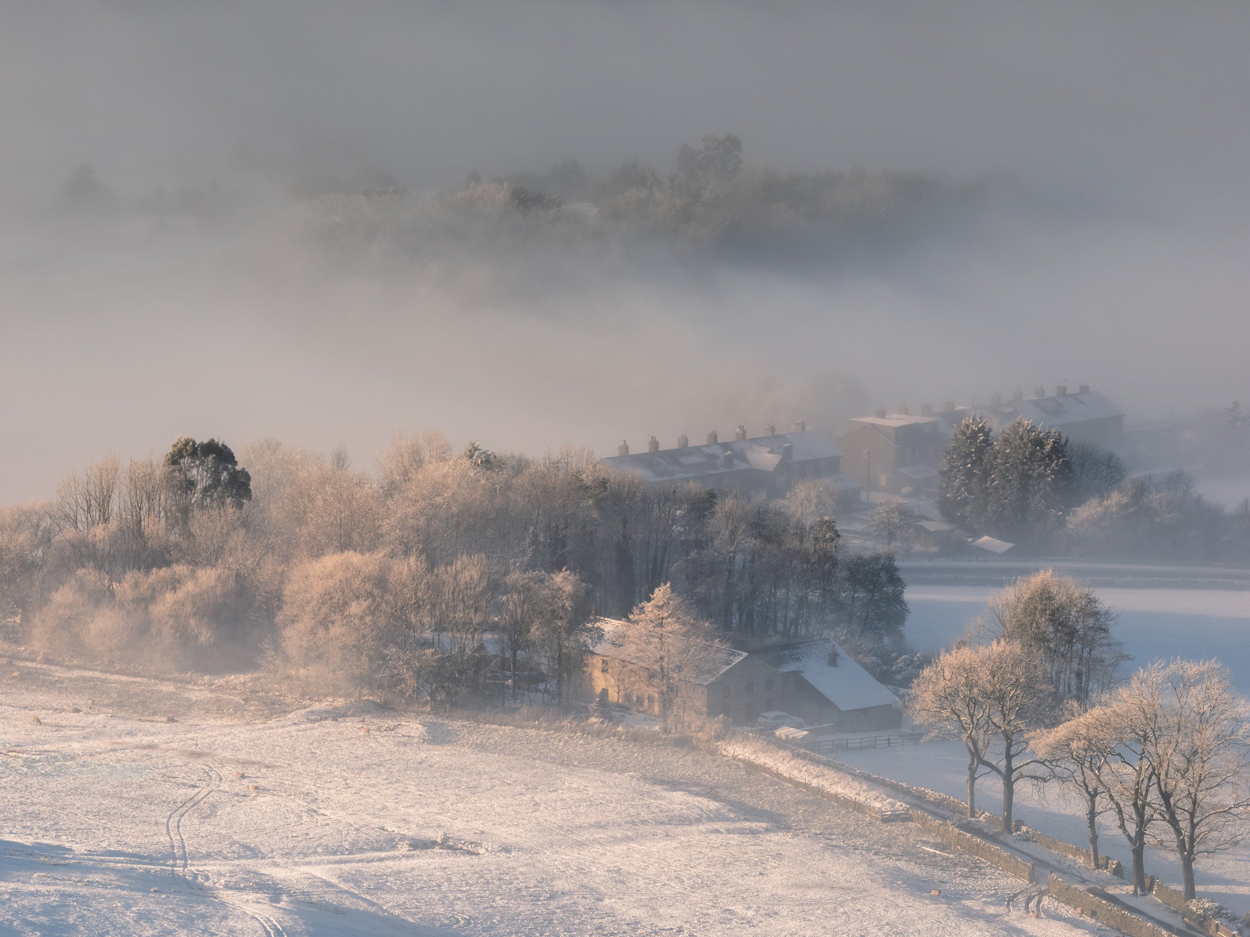 Snow-covered stone farm buildings and frosted trees in Newgate Valley, Rossendale, lit by warm morning sunlight beneath a layer of low mist.