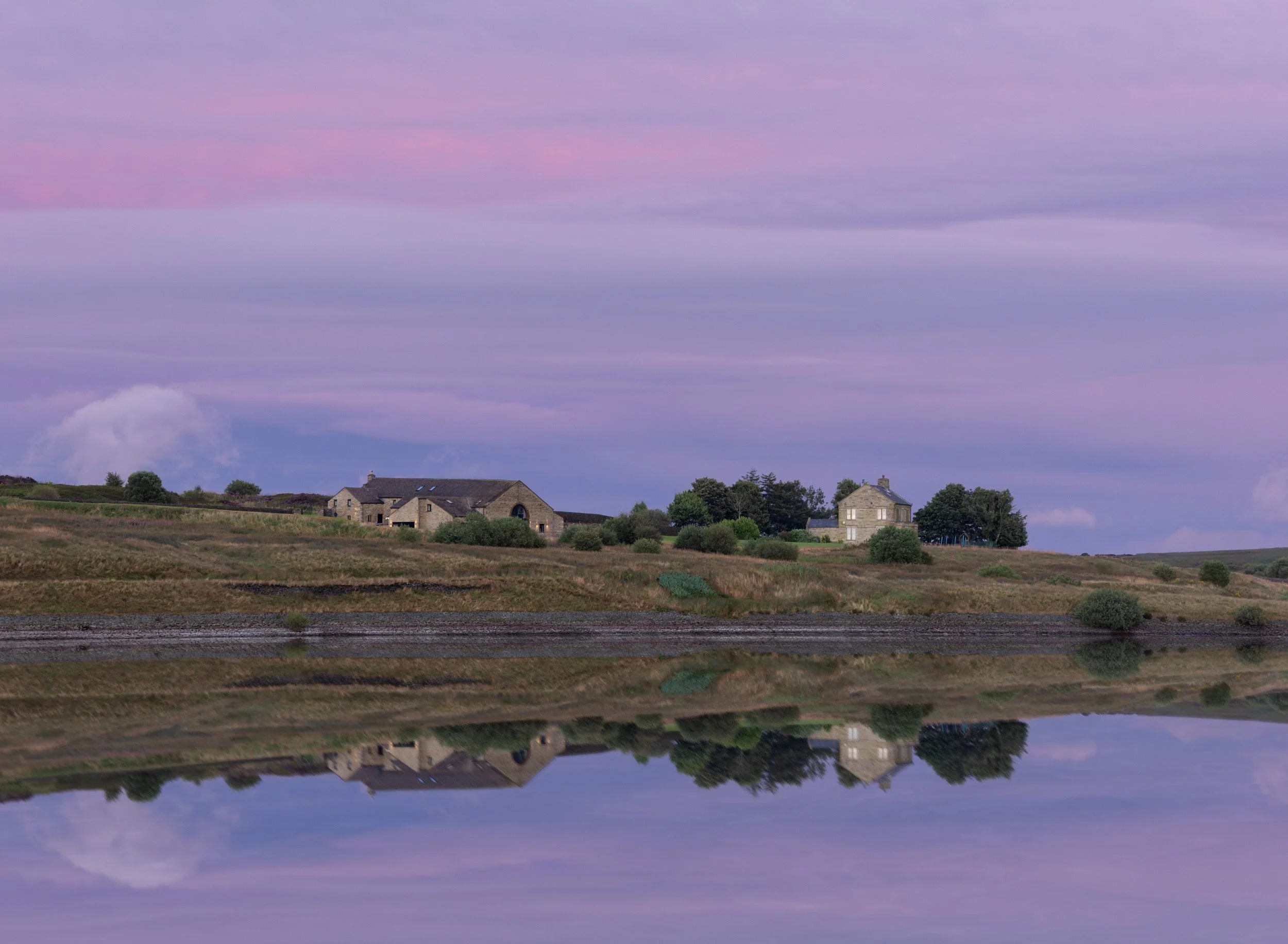 Ashworth Moor Reservoir in Norden near Rochdale, reflecting stone farm buildings and a purple dusk sky on its calm, glassy surface.