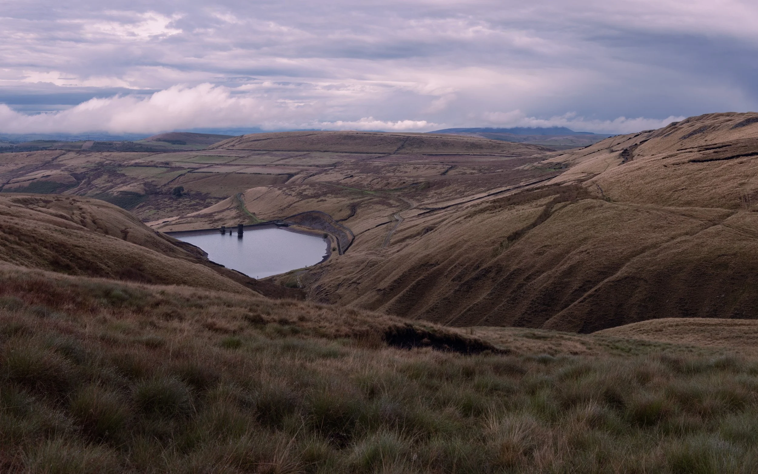 Low white clouds drifting along distant hills above the water of Scout Moor High Level Reservoir.