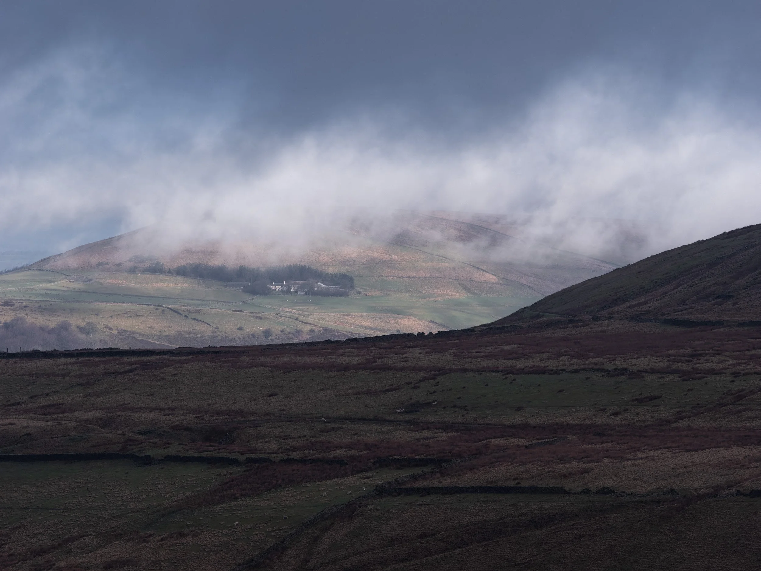Telephoto landscape shot of low-hanging white clouds drifting over a sunlit hillside farm framed by shadowed moorland foreground.