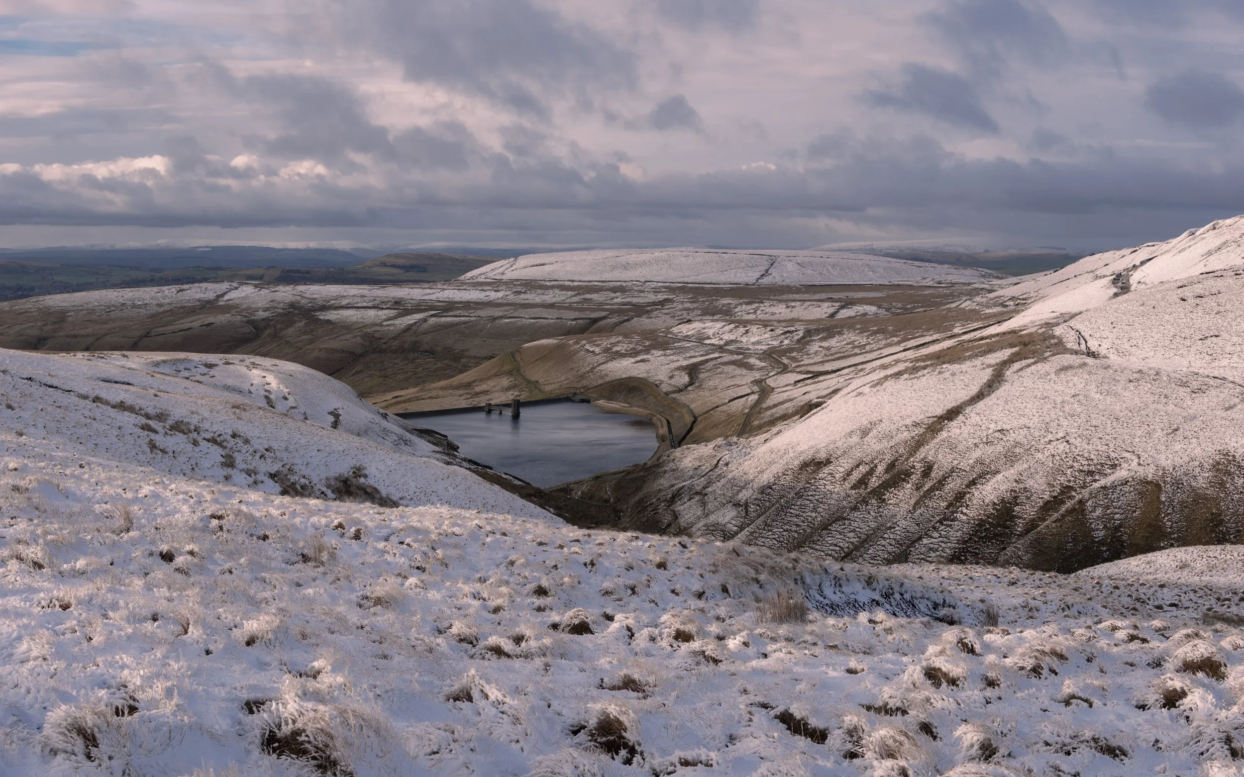 Wide elevated view across snow-dusted moorland slopes toward Scout Moor High Level Reservoir under a moody, clouded sky