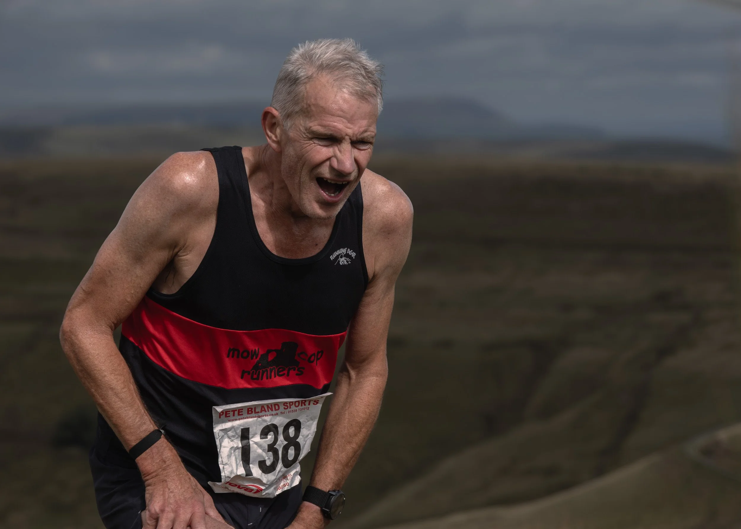 Close-up of runner #138 in a black and red Mow Cop Runners vest grimacing with open-mouthed effort on the Scout Moor fell race.