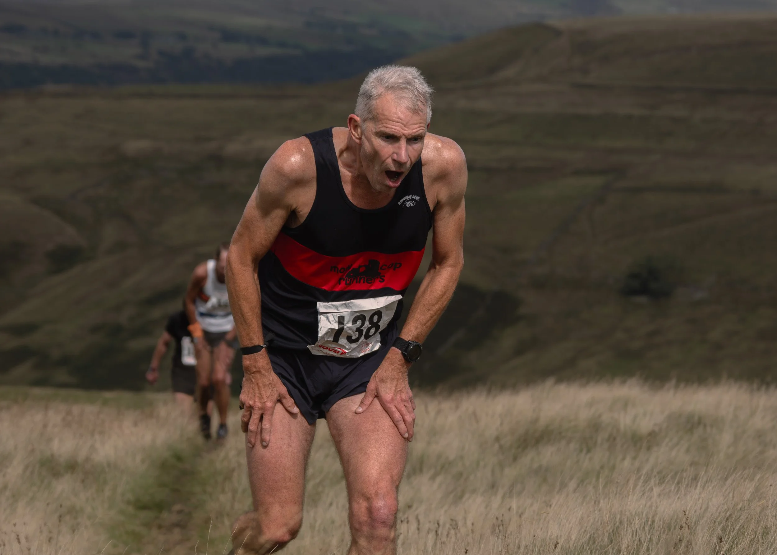 Fell runner wearing race number 138 from Mow Cop Runners leaning forward with hands on knees during a steep climb on Scout Moor.
