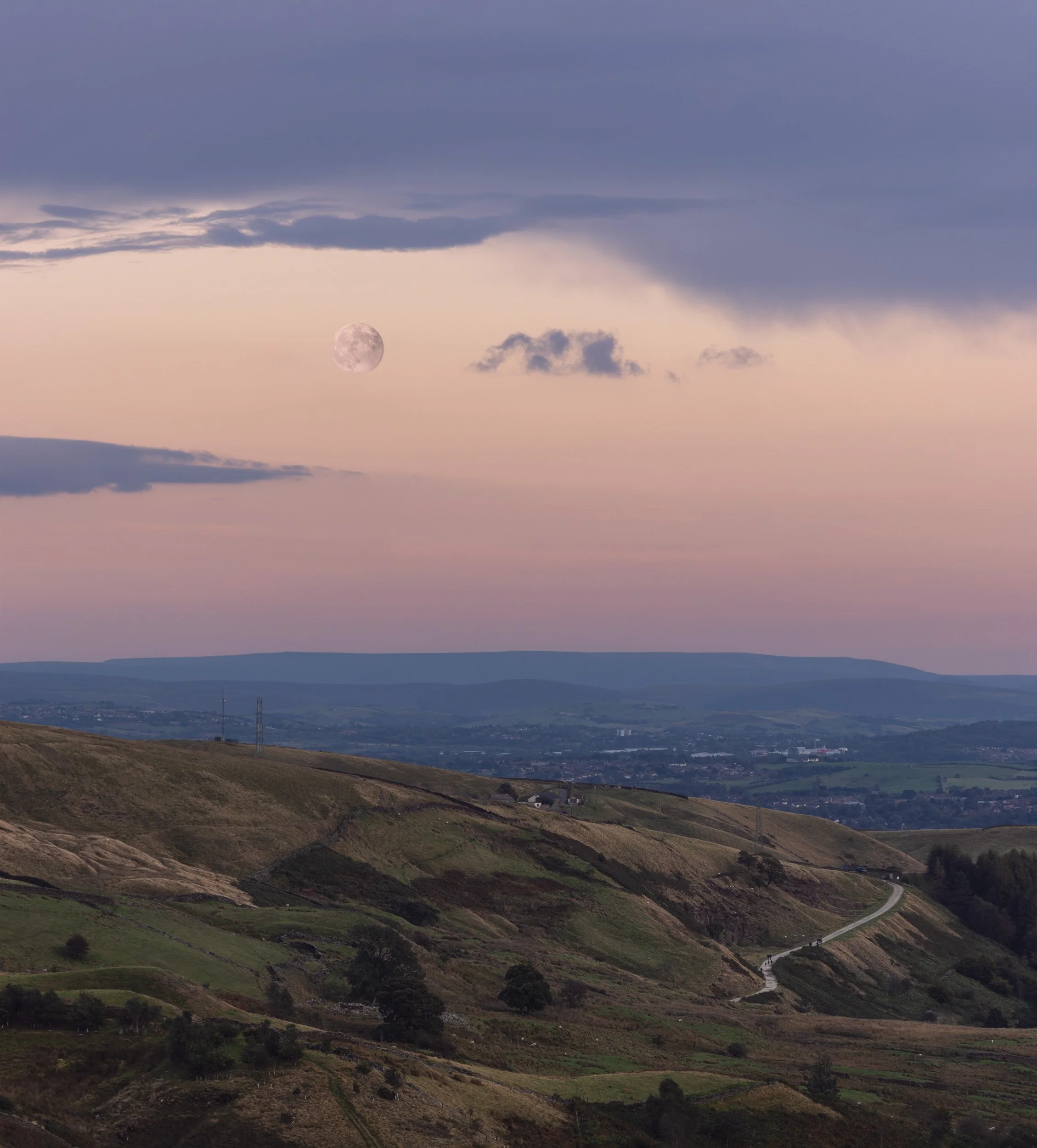 Full moon rising over the grassy ridges and rolling hills of Naden Valley near Scout Moor, Norden, under a pale pink dusk sky.