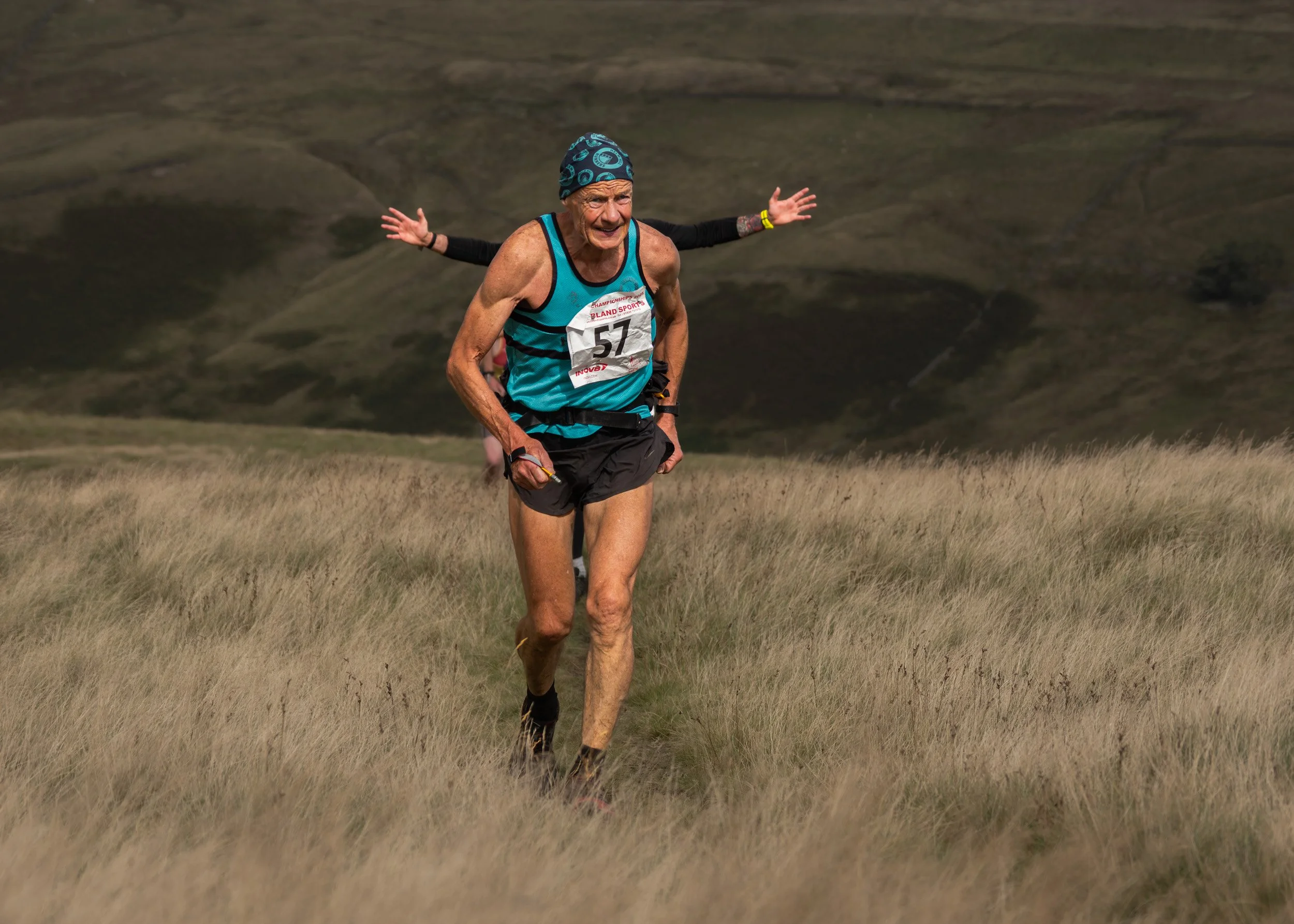 Veteran fell runner wearing race number 57 and a blue headband charging uphill on Scout Moor while another runner stretches her arms out behind him.