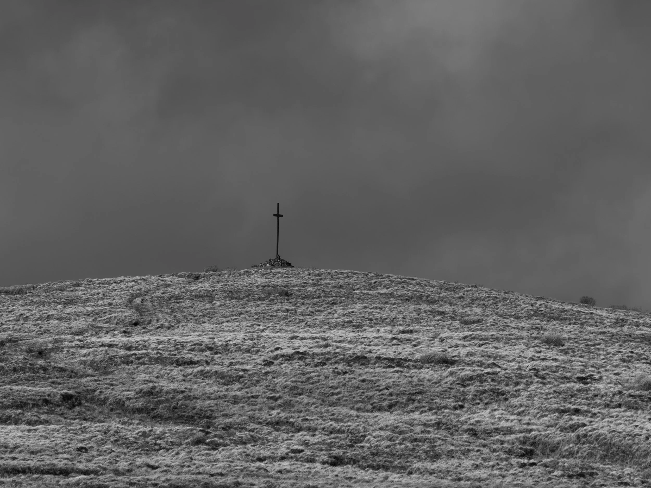 Monochrome photograph of the summit cross on Whittle Pike standing against a dark, brooding storm sky on Scout Moor.