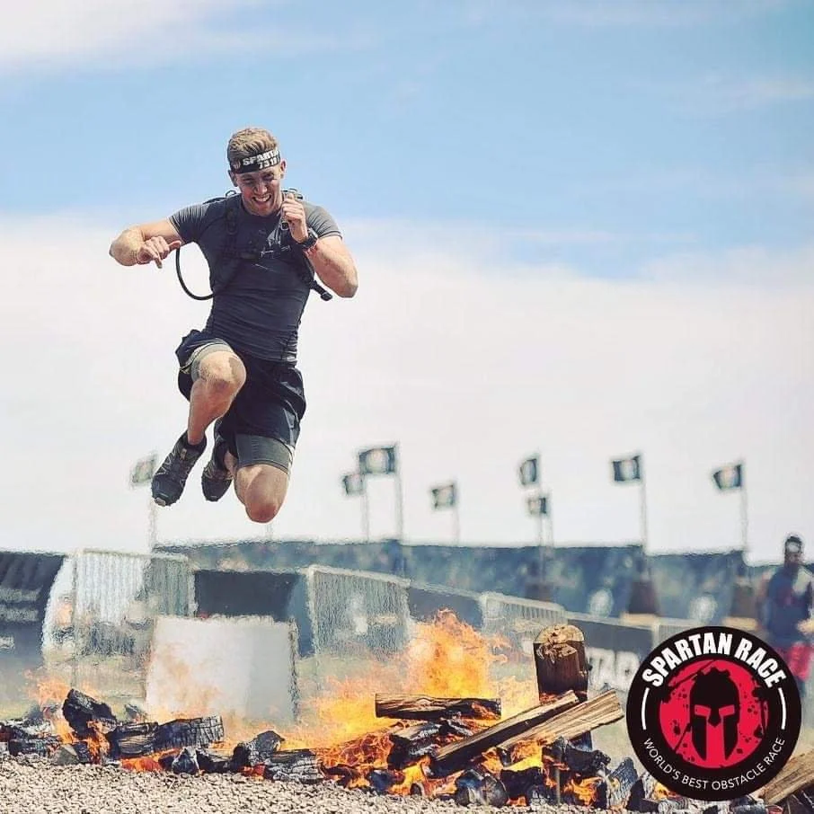 A man dressed in athletic clothes jumping over a line of burning logs and rocks during an obstacle race, with a Spartans Race logo in the bottom right corner.