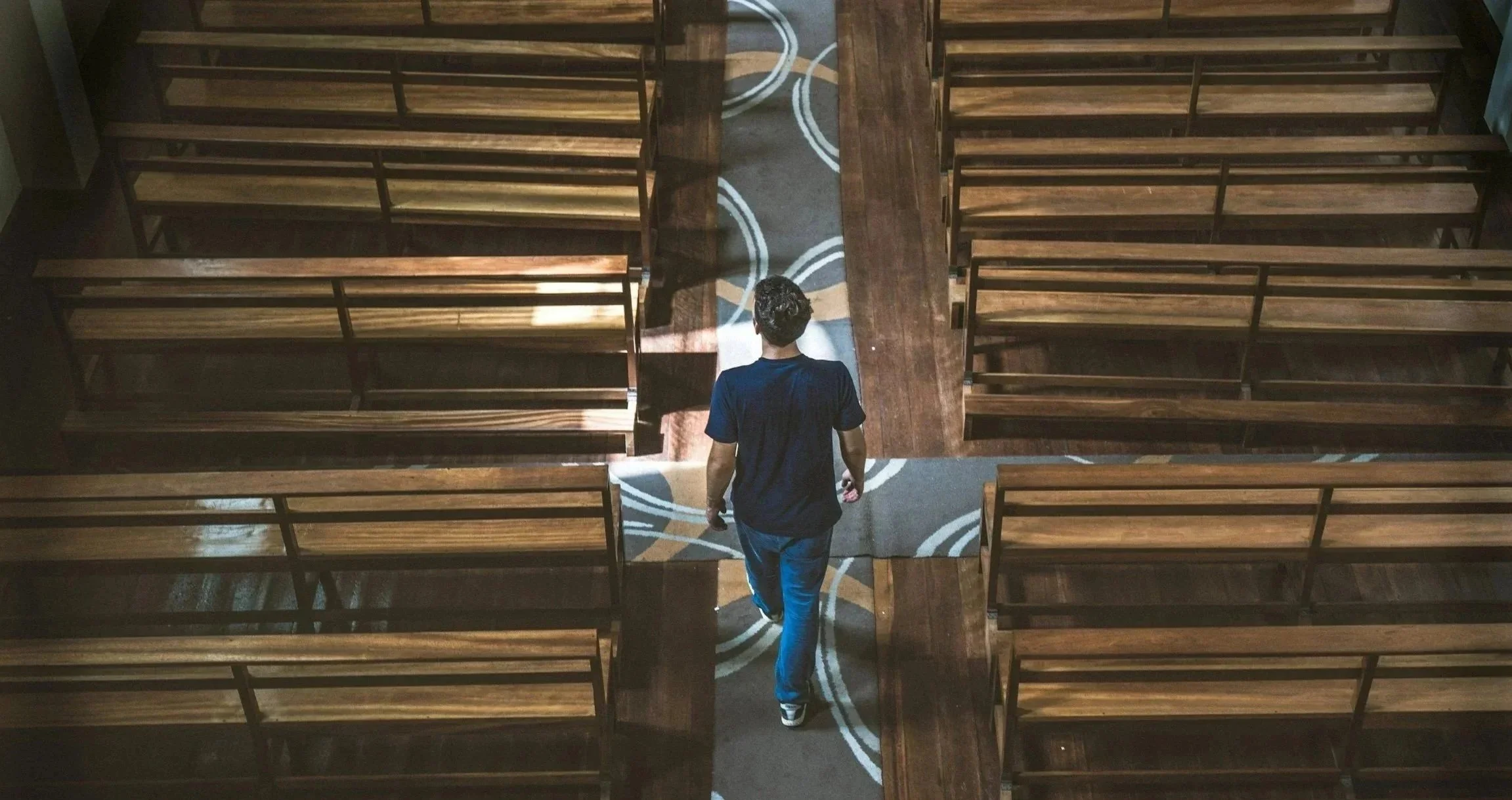 A person walking down an aisle between empty wooden pews in a church or auditorium. The person is viewed from the back, wearing a dark blue shirt and jeans, and the aisle is decorated with a circular pattern on the carpet.