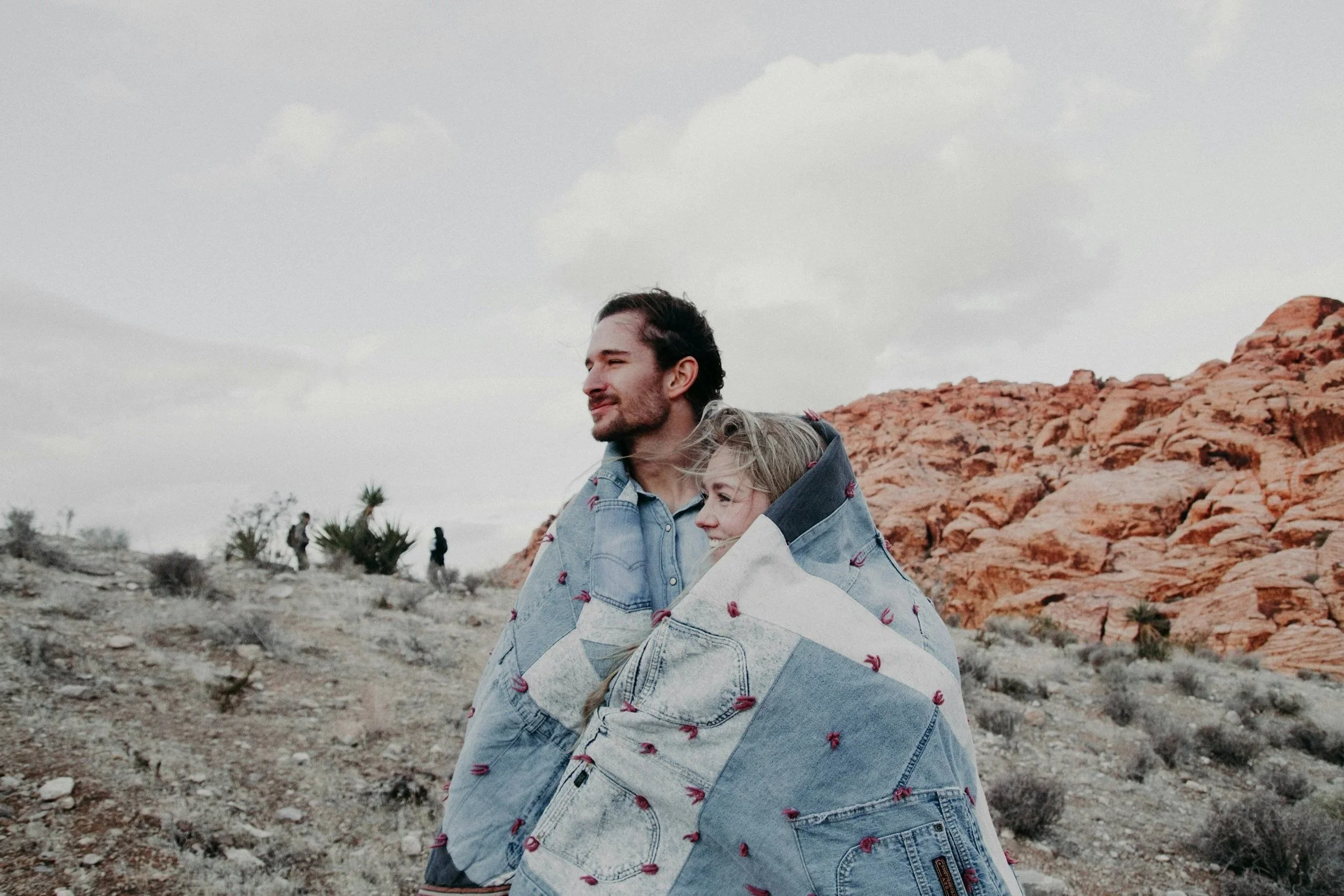 Two people, a man and a woman, sitting outdoors in a desert landscape with red rocks and sparse vegetation, wrapped together in a denim blanket, smiling and looking into the distance under a cloudy sky.