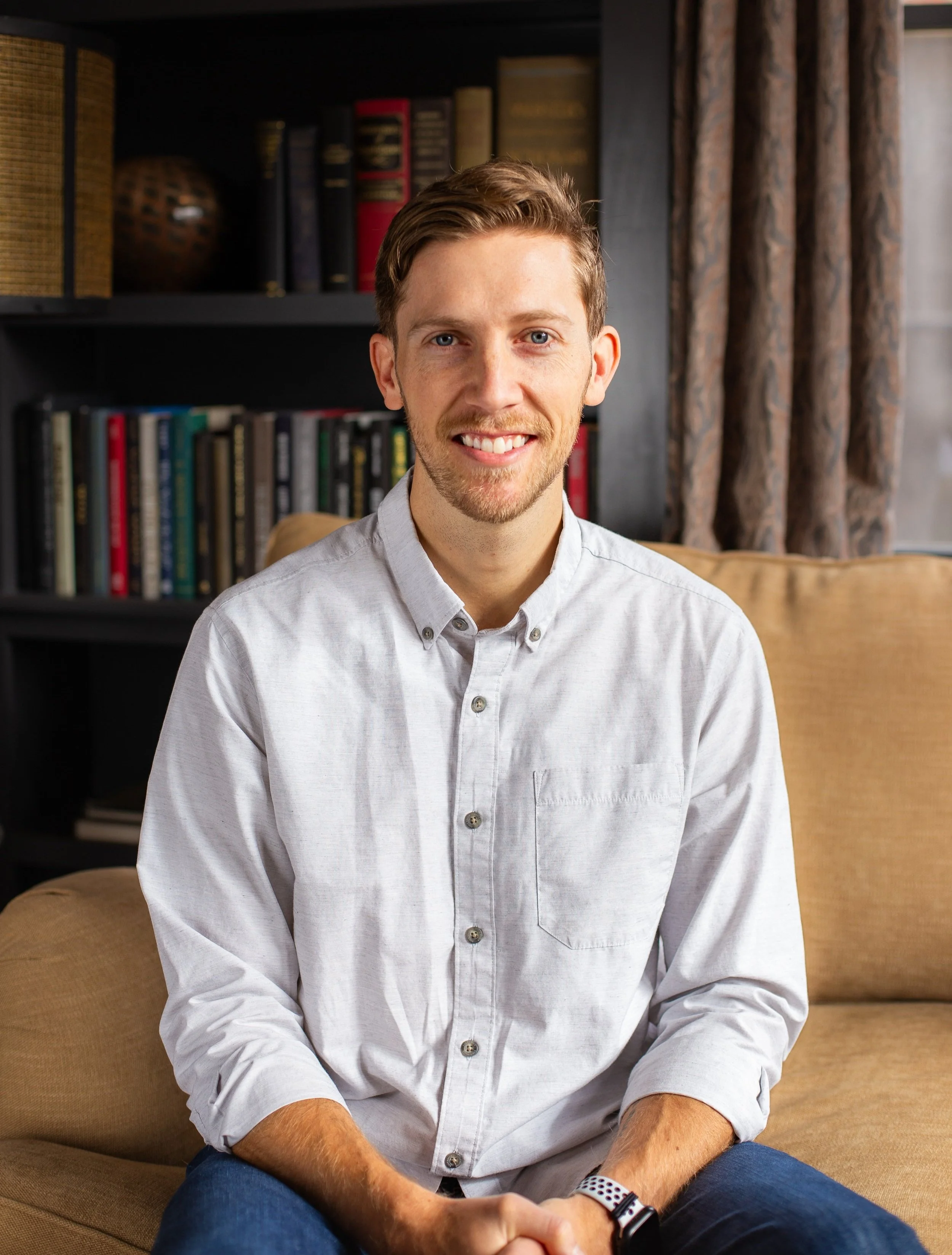 A young man with light brown hair and blue eyes smiling while sitting on a beige couch in a living room with a black bookshelf filled with books behind him and a brown curtain on the window.