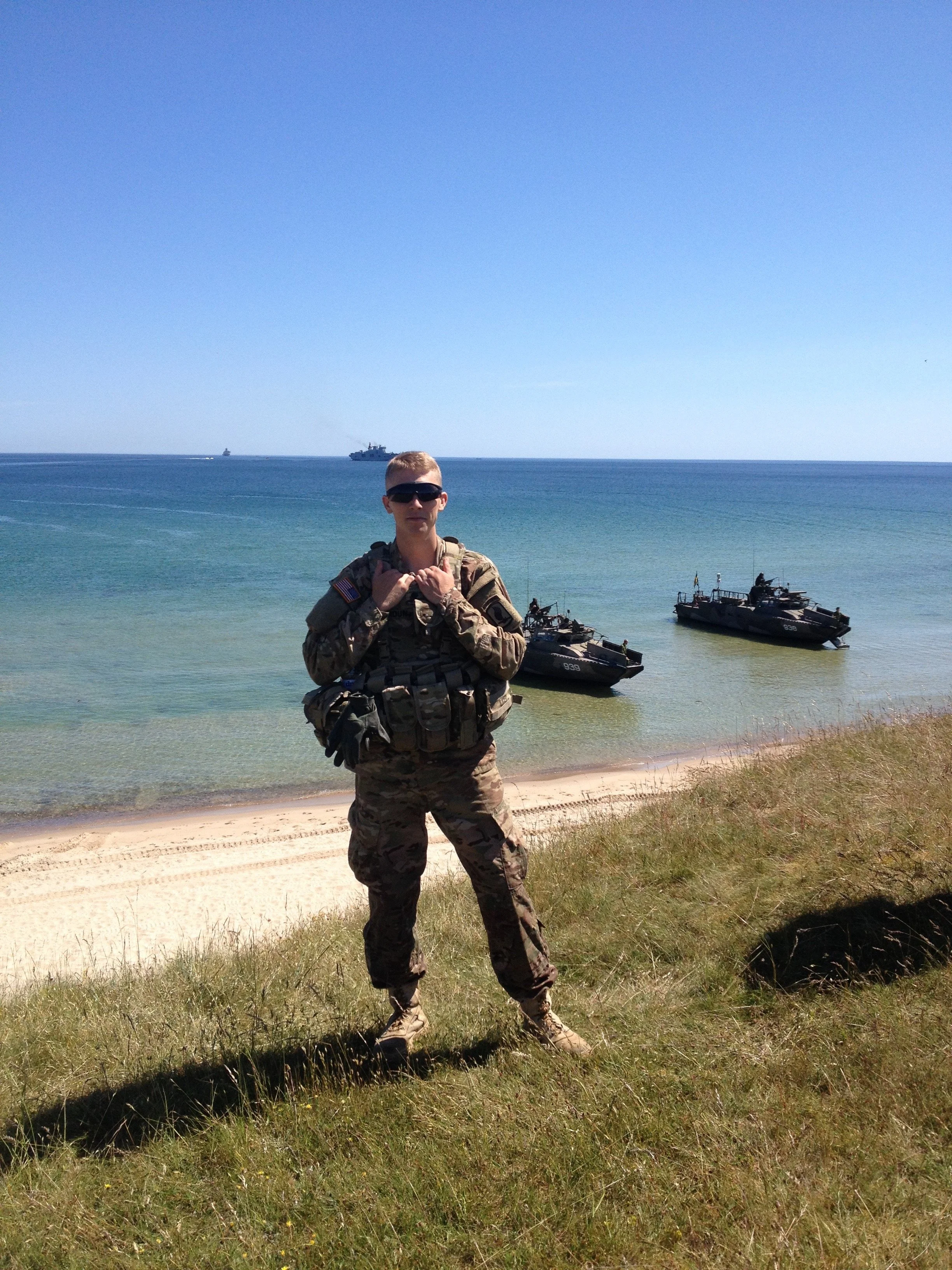 A soldier in camouflage uniform and sunglasses stands on a grassy shoreline with military boats and ships in the water behind him under a clear blue sky.