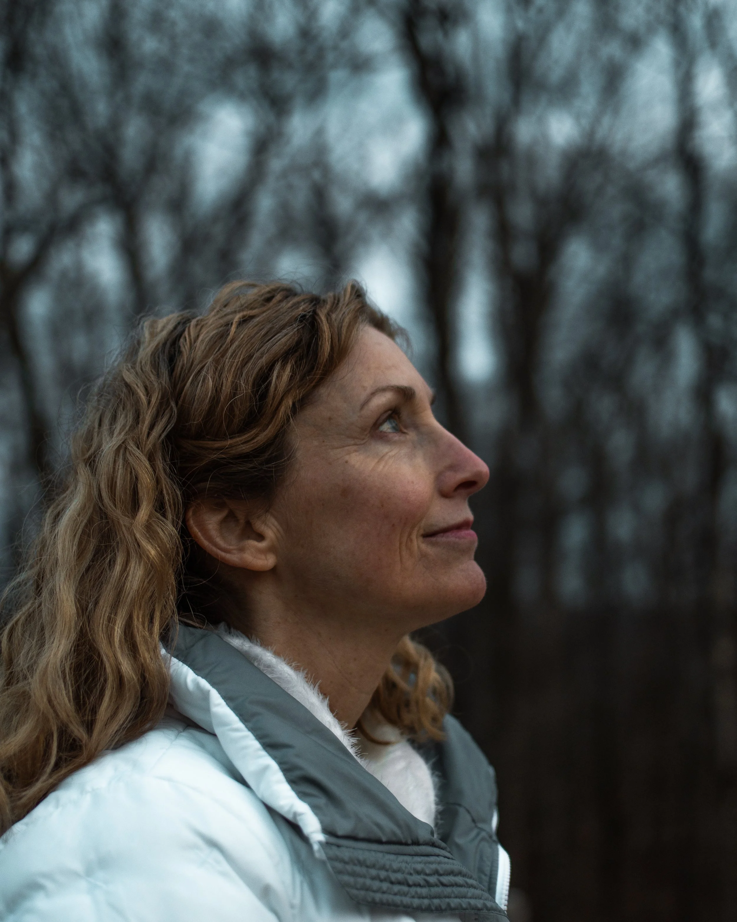 Side profile of a woman with red curly hair outdoors, wearing a light-colored jacket, with blurred trees in the background.