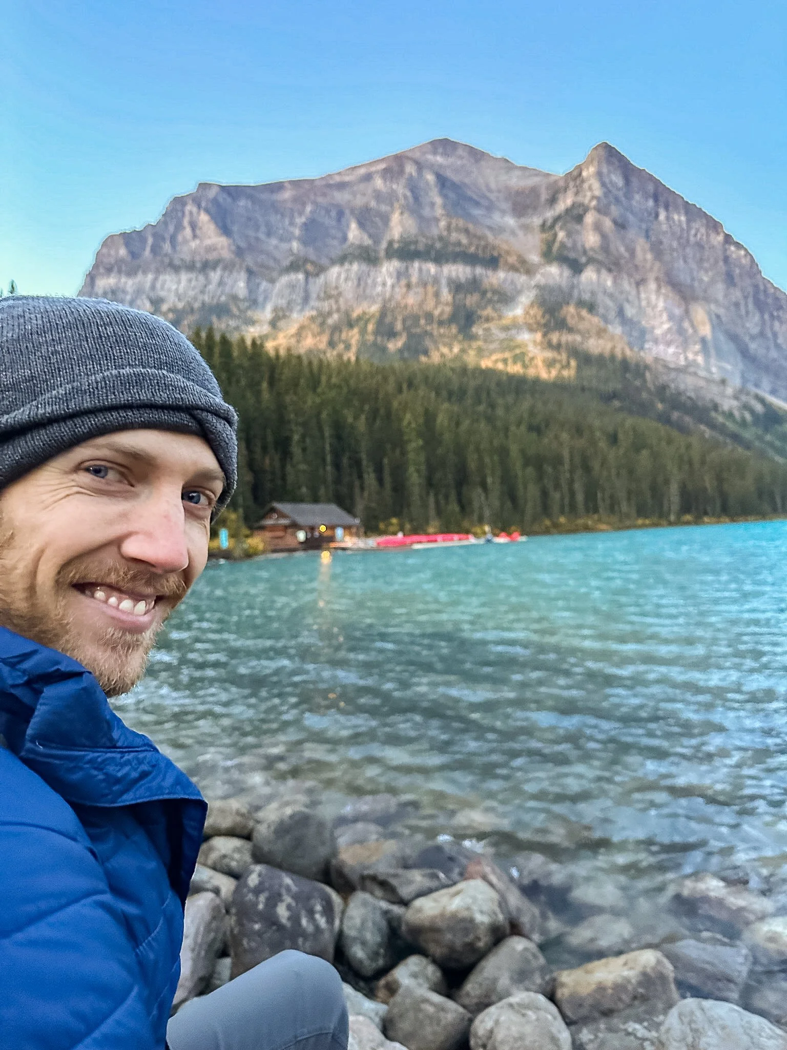 A man smiling, wearing a gray beanie and a blue jacket, sitting on rocks by a lake with a mountain in the background.
