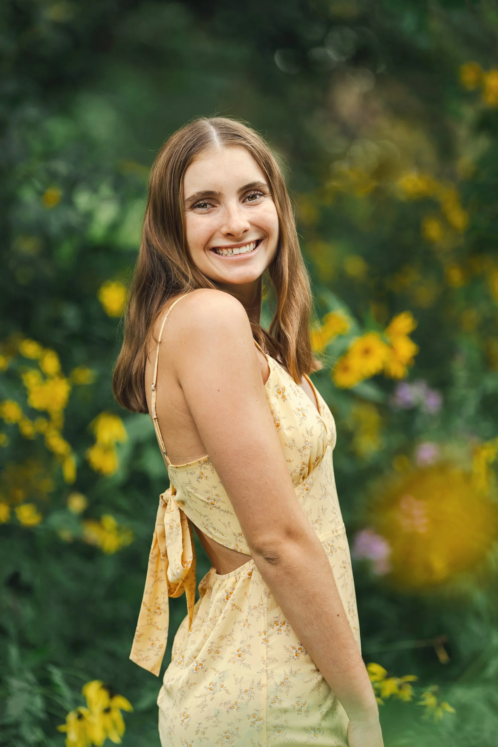 A young woman with brown hair smiling outdoors, wearing a yellow floral dress with cut-out sides, surrounded by green foliage and yellow flowers at the McCrory Gardens in Brookings, South Dakota for her high school senior photos.