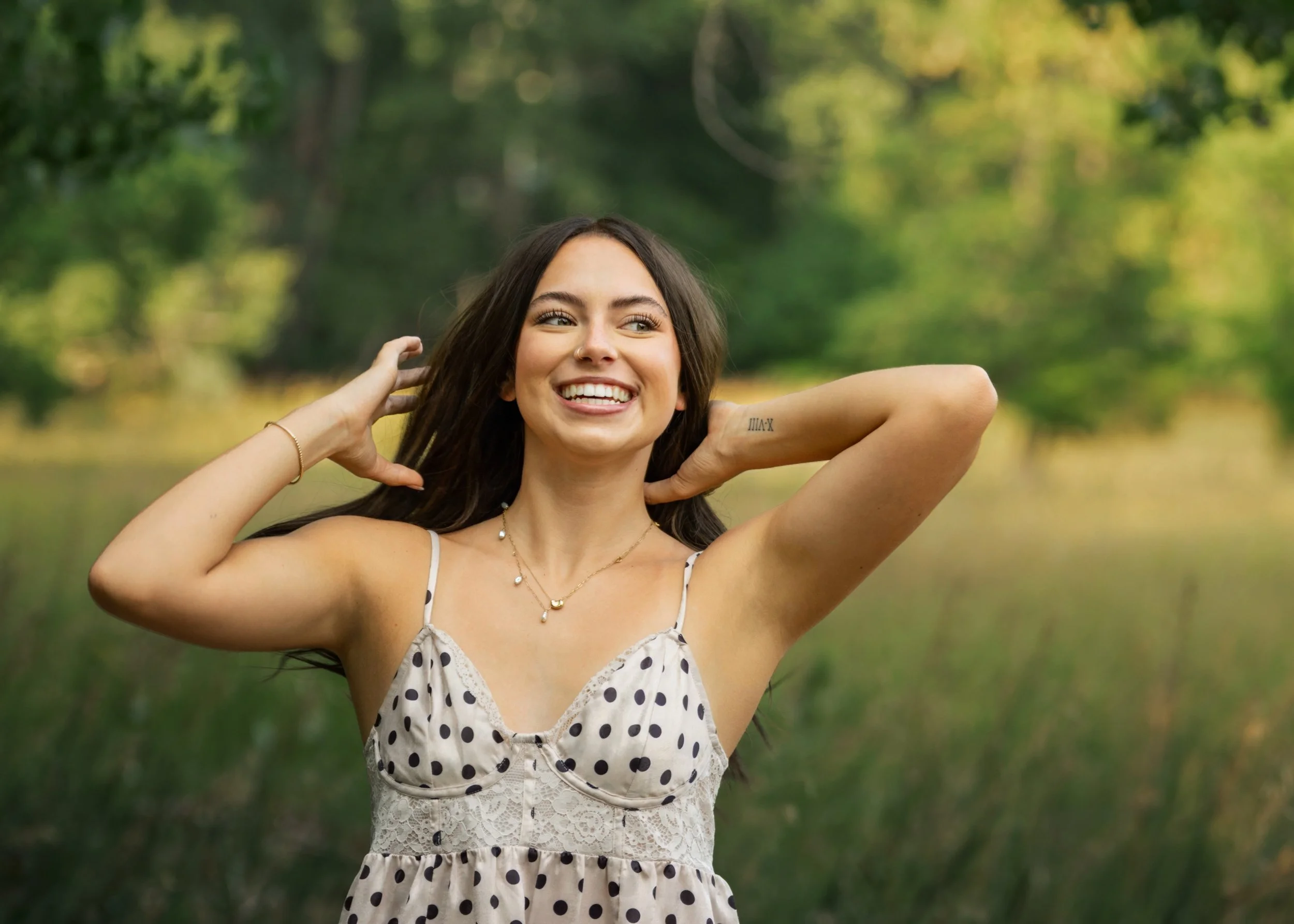 A high school senior girl pulls her hair back while laughing in a white polka dot dress in Fort Collins, Colorado.