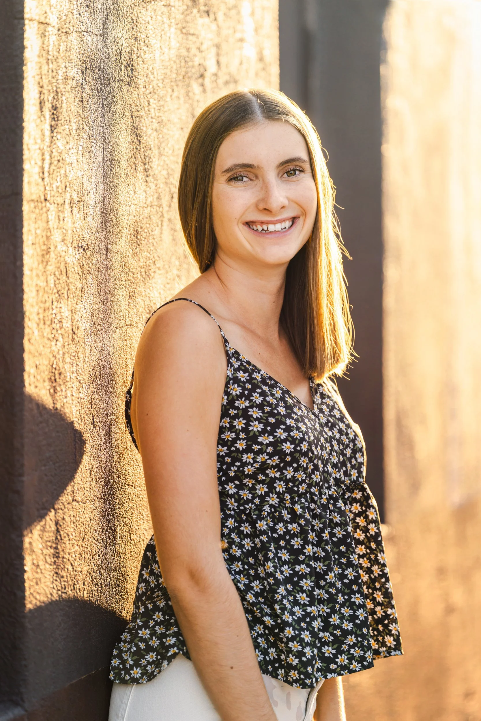 A young woman with long brown hair, wearing a black floral tank top, smiling and standing against a textured wall in sunlight in Buffalo, Wyoming for her northern Colorado senior portraits.