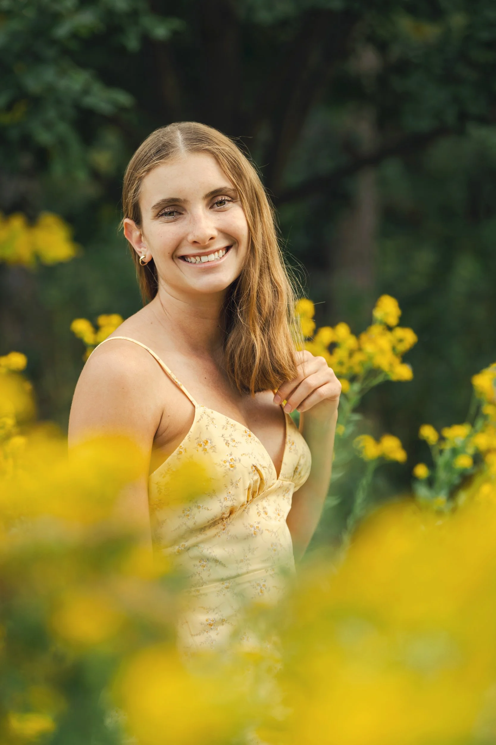 A young woman with auburn hair wearing a yellow floral dress, smiling in the McCrory Gardens (Brookings, South Dakota),  with yellow flowers and green foliage for her high school senior portraits.