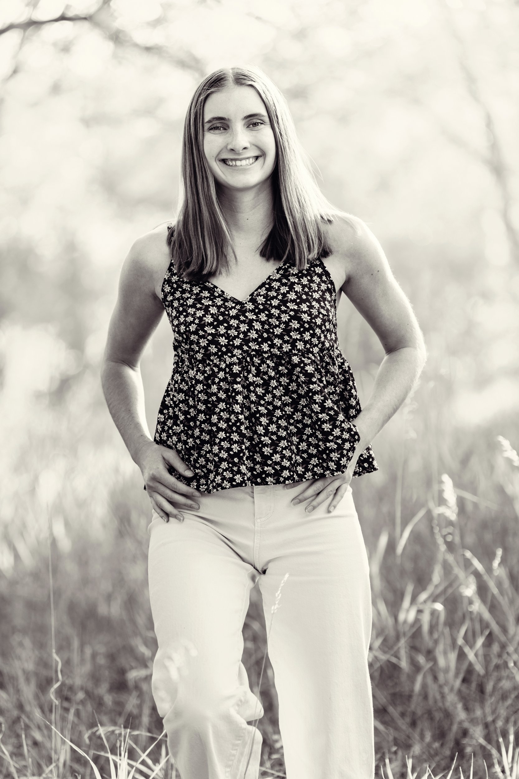In a black and white shot a Liberty Common high school senior woman walks towards the camera smiling in white jeans and a black top in a Buffalo, Wyoming natural area with soft glowing light surrounding her.