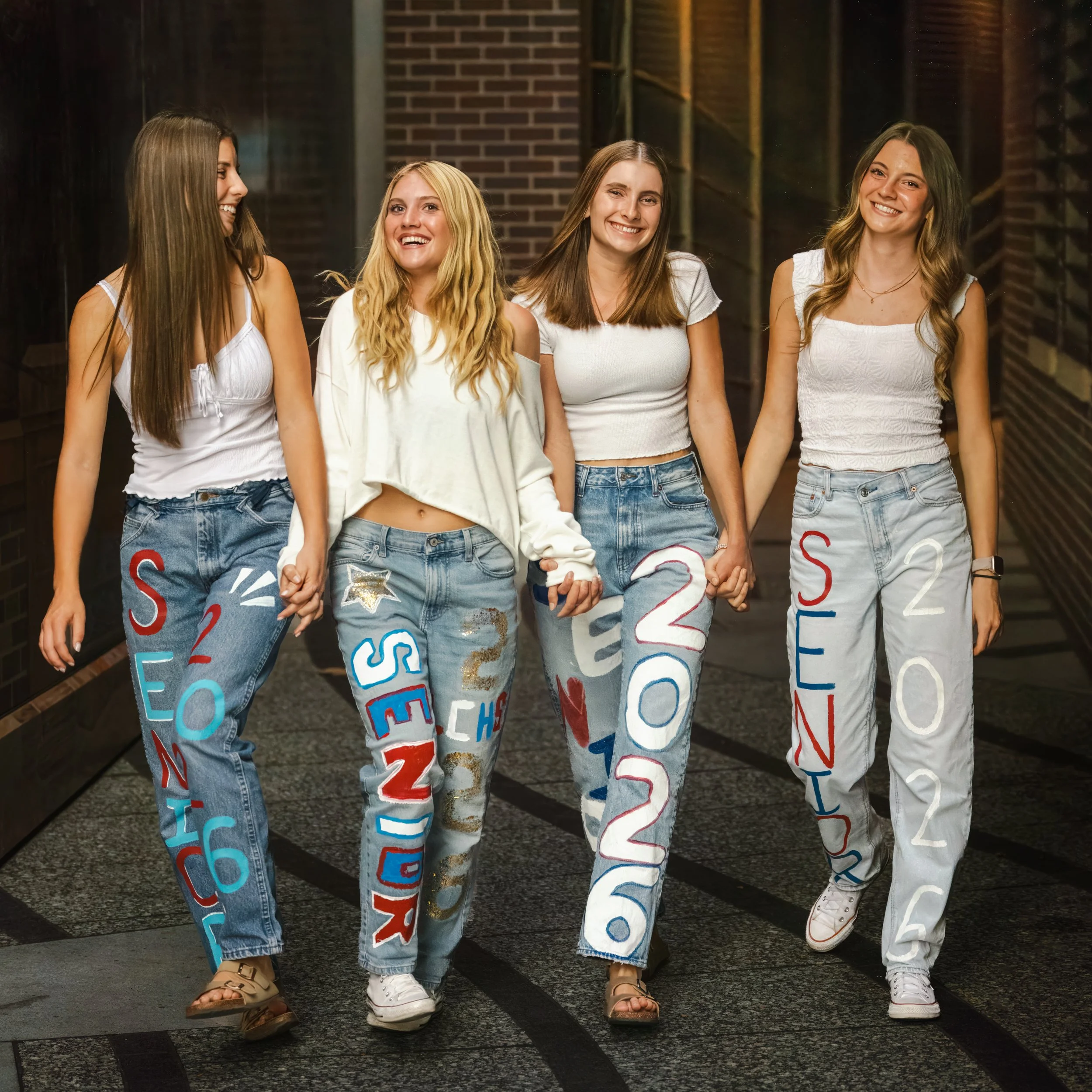 Four young women (high school seniors) wearing jeans with colorful letters and numbers, holding hands, walking and smiling in an urban setting in Fort Collins, Colorado.