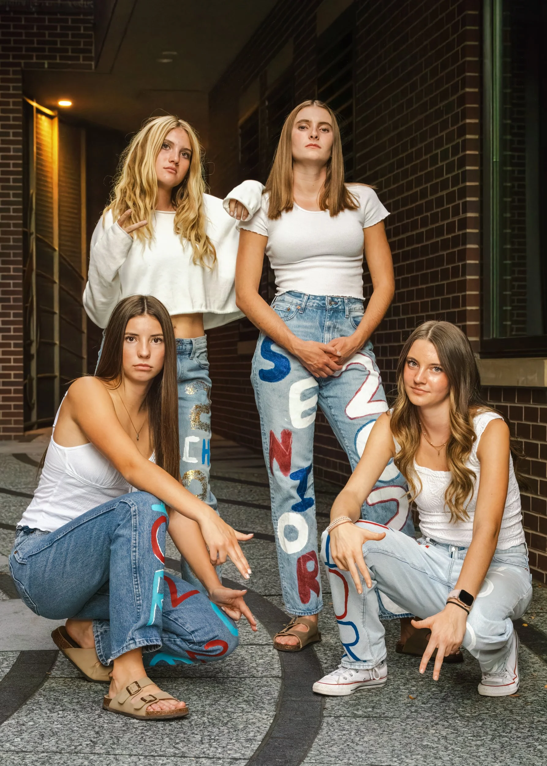 Four young women (high school friends) posing together outdoors in an alley a la Charlies Angels, wearing casual outfits with painted, colorful letters on their jeans and tops in Fort Collins, Colorado.