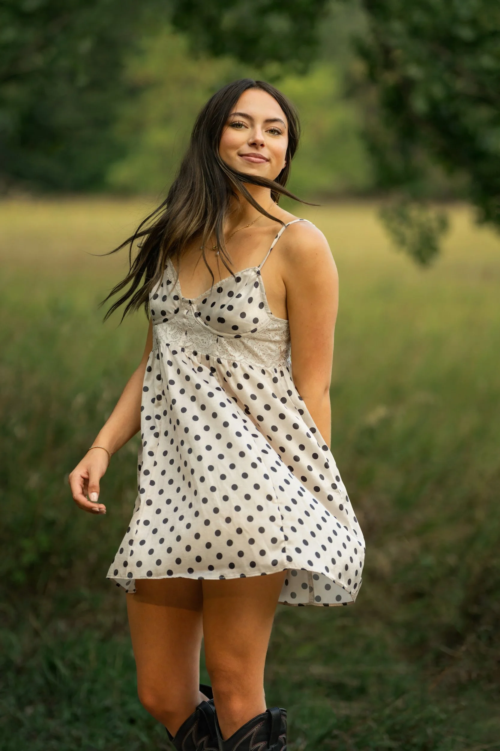 A young woman with long dark hair in a white spaghetti strap polka dot dress standing outdoors in a grassy area with trees in the background for her high school senior portraits in Ft. Collins, Colorado.