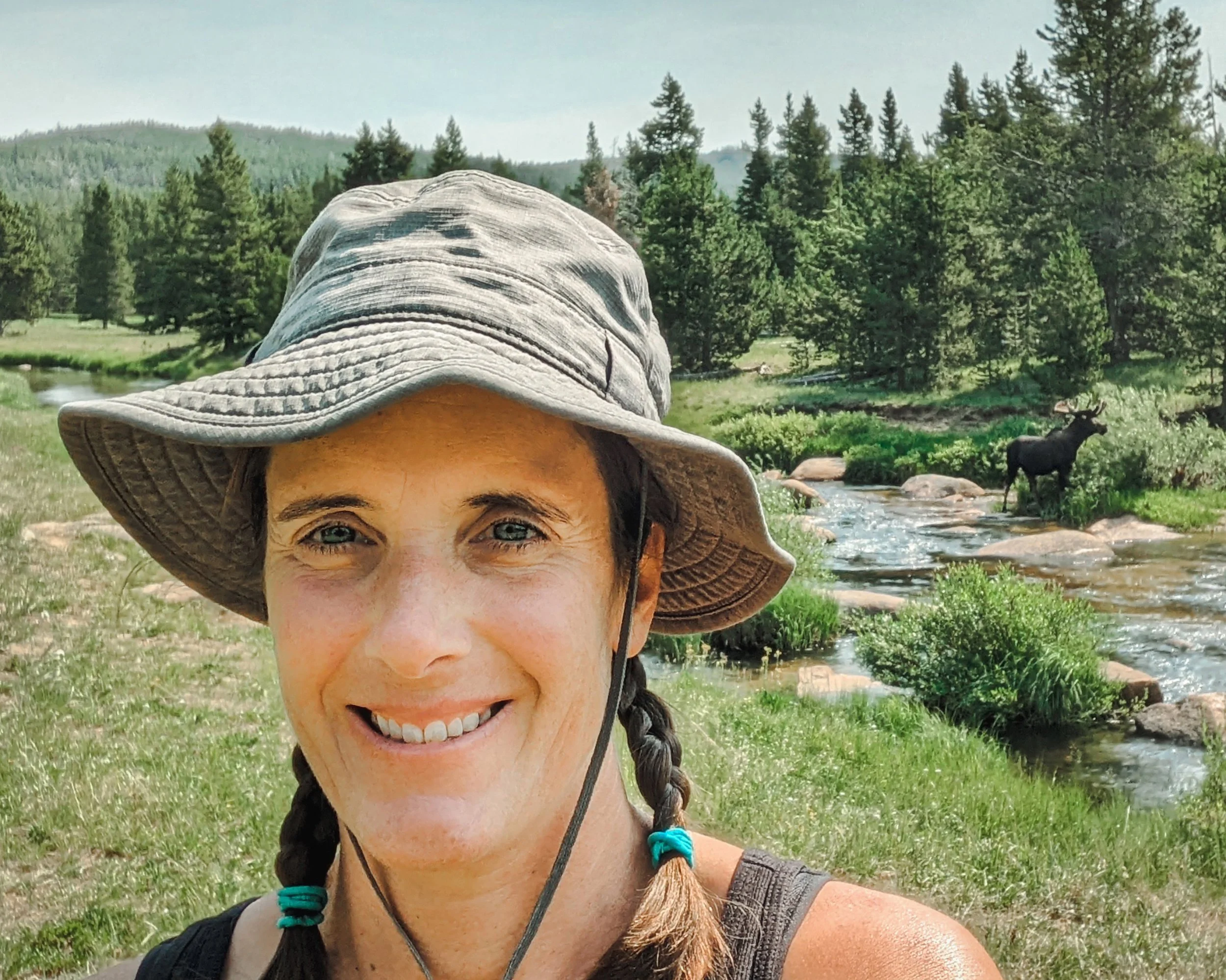 Jess Barr smiling at a camera while a moose munches on willows behind her