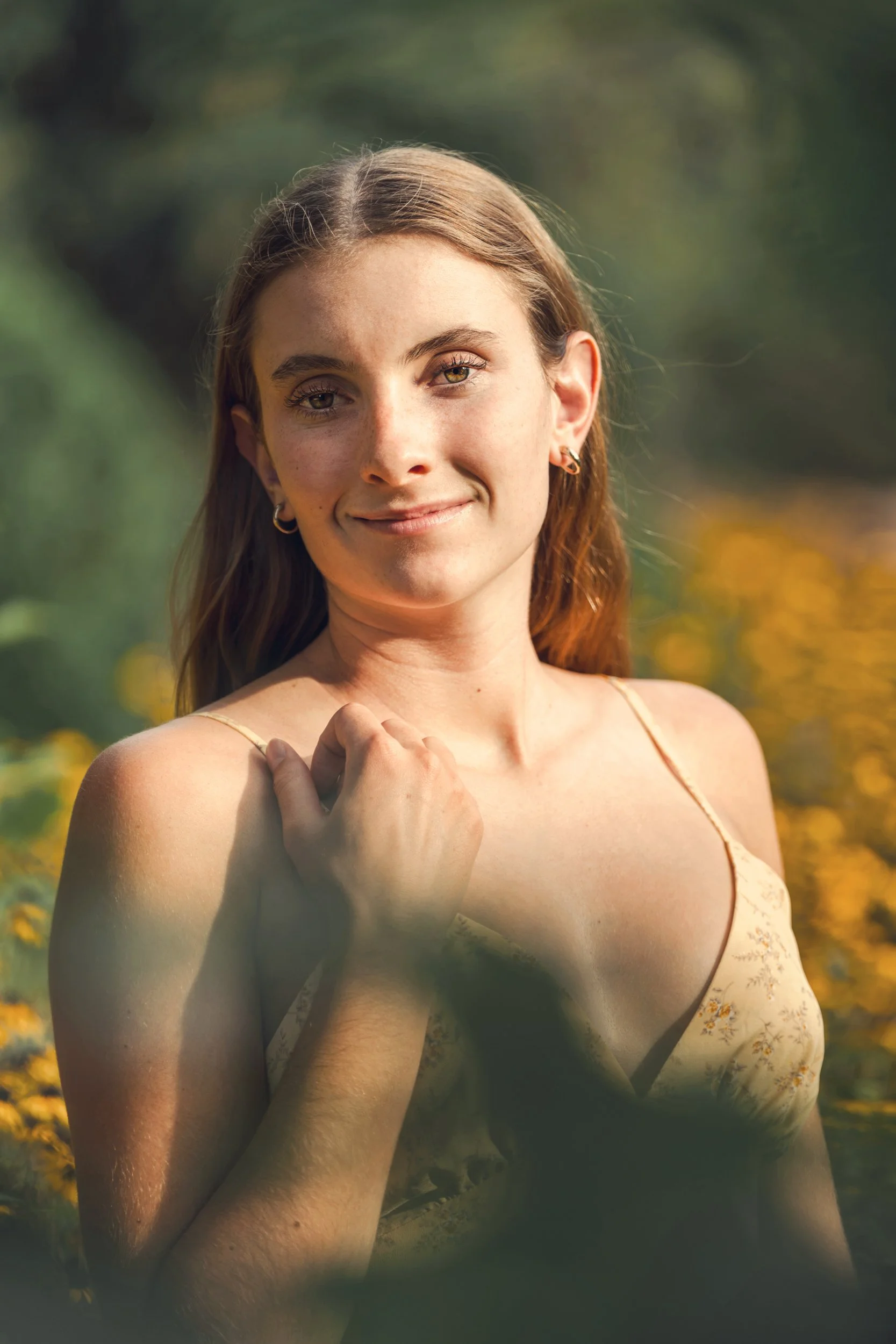 A young woman with long brown hair and light skin standing outdoors in a natural setting with yellow flowers and green foliage, wearing a floral dress, smiling softly at the camera with one hand near her shoulder.