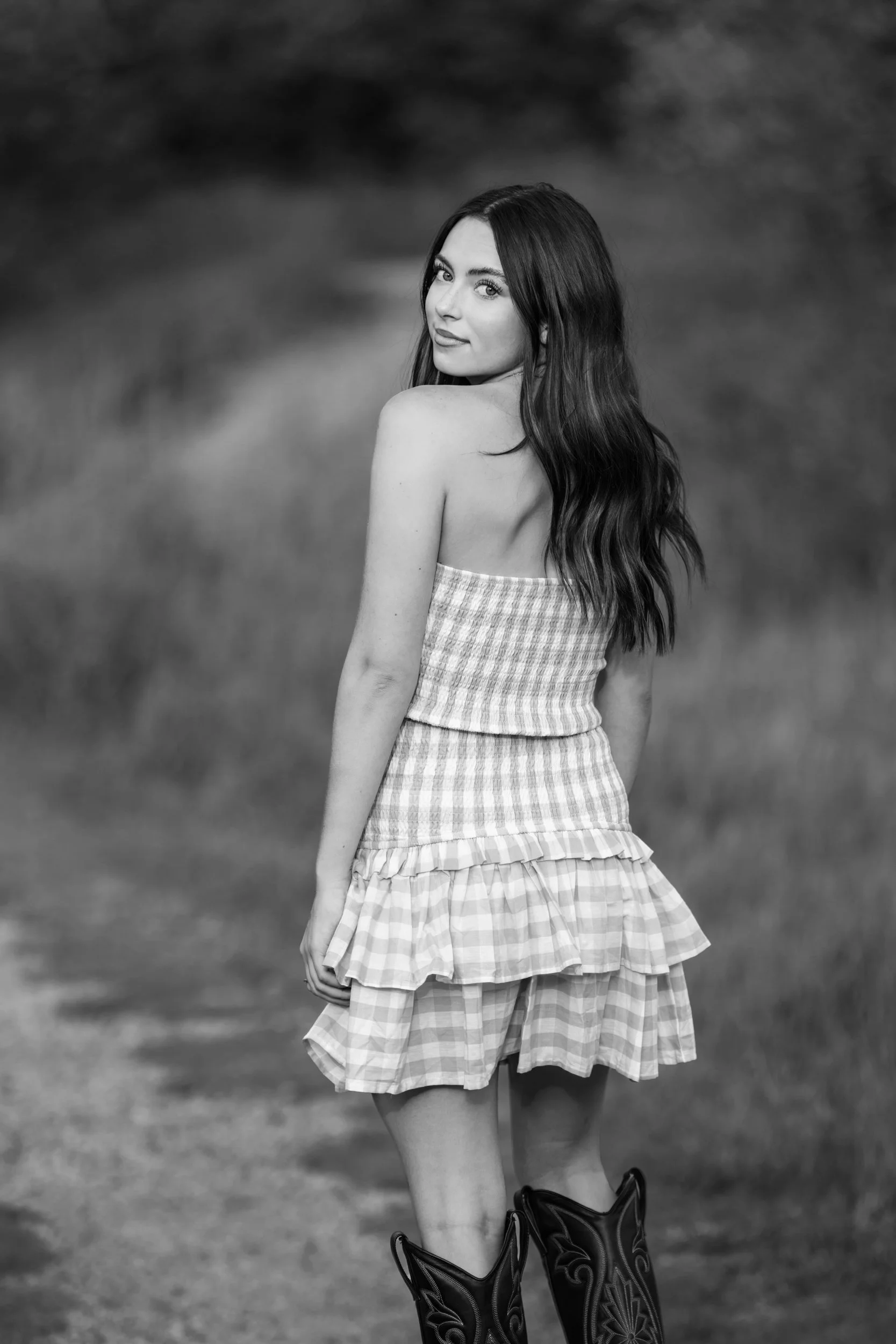A woman with long, wavy hair stands outdoors on a dirt path, turning slightly to her left and gazing at the camera. She is wearing a white, strapless checkered top and a layered, checkered skirt, with cowboy boots. 