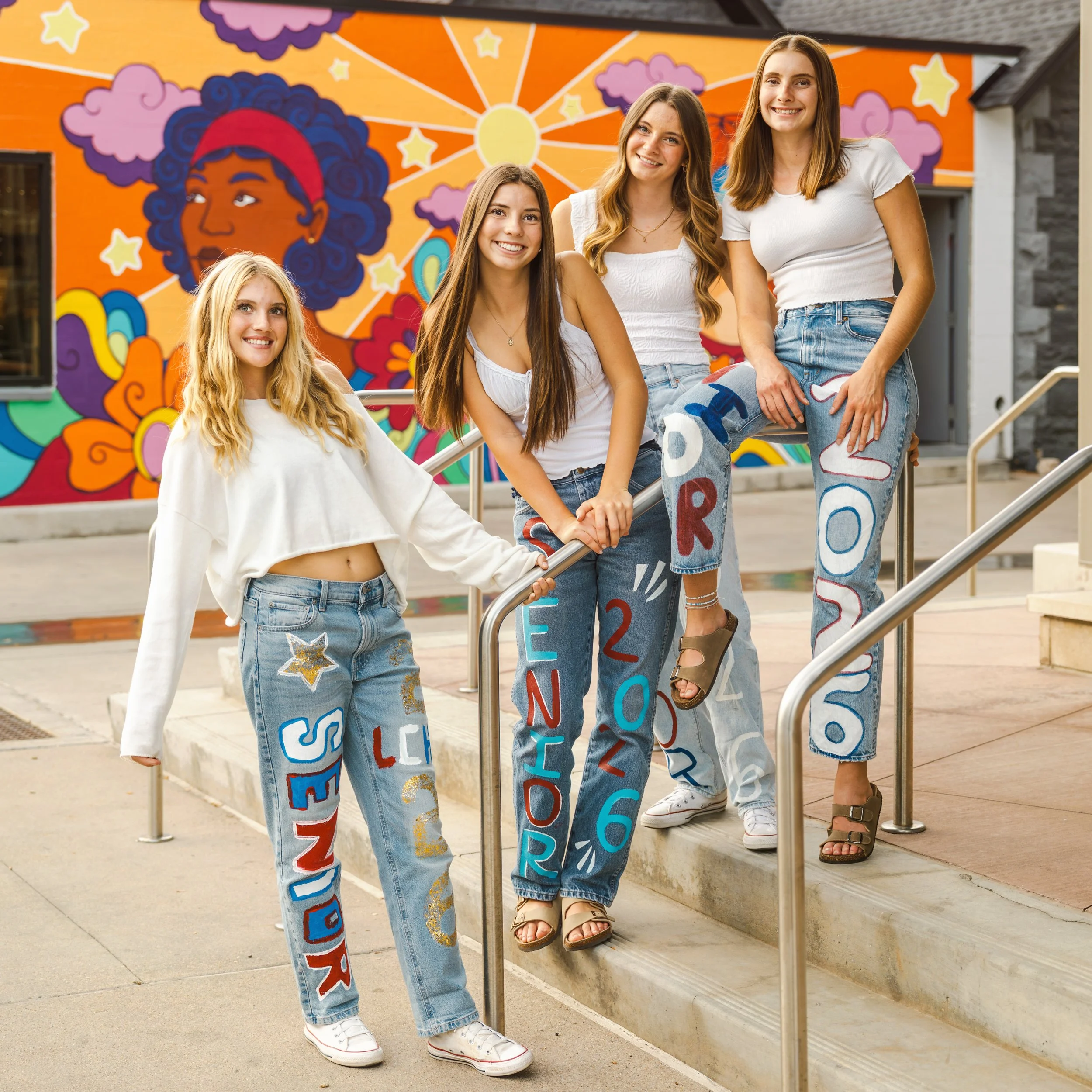 Four high school senior girlfriends standing on stairs with a colorful mural near The Elizabeth Hotel in downtown Fort Collins, Colorado in the background, celebrating graduation with custom decorated jeans featuring their graduation year.