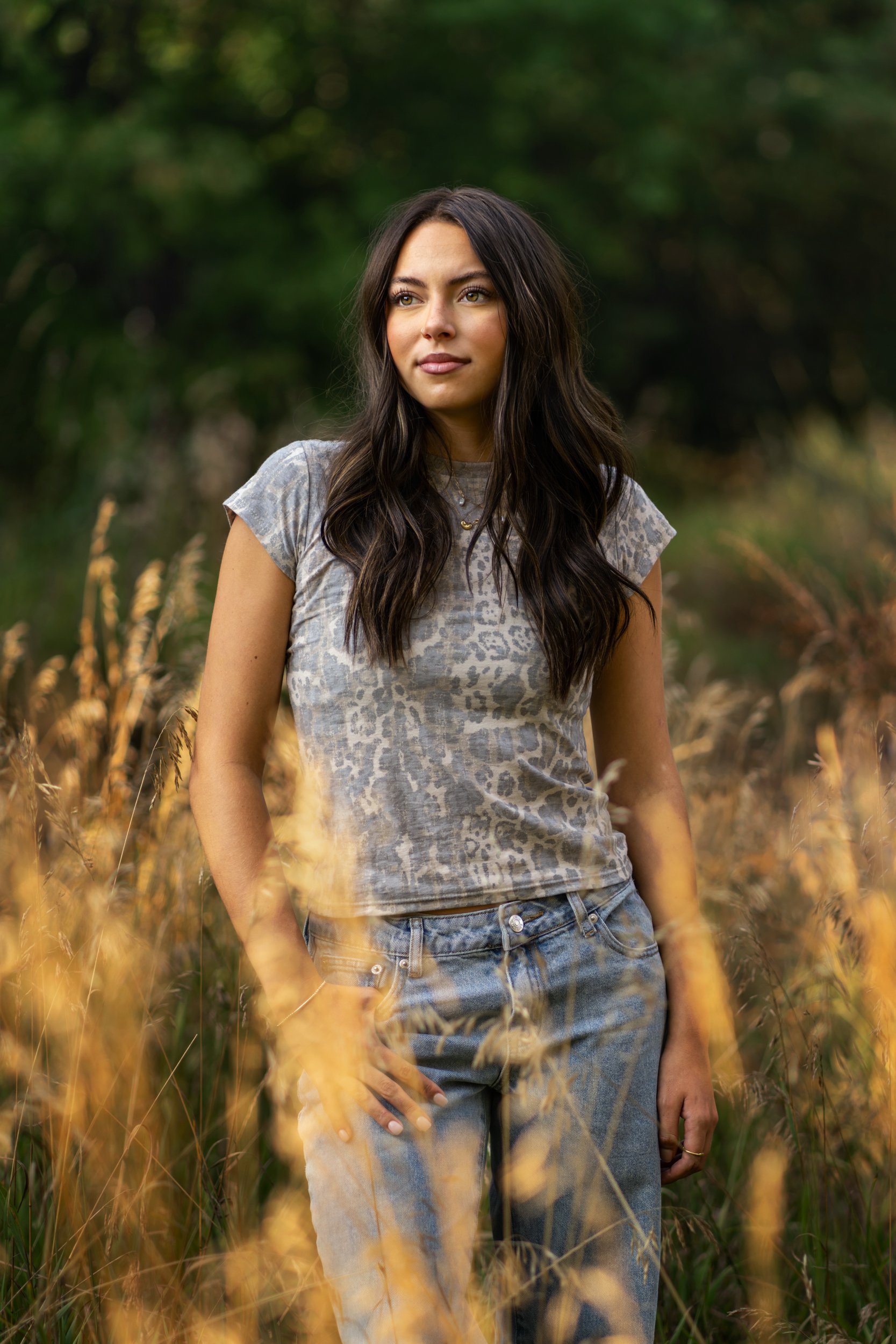 A young woman with long dark hair standing in a field of tall, golden grass with a blurred green trees background in Ft. Collins, Colorado natural area.