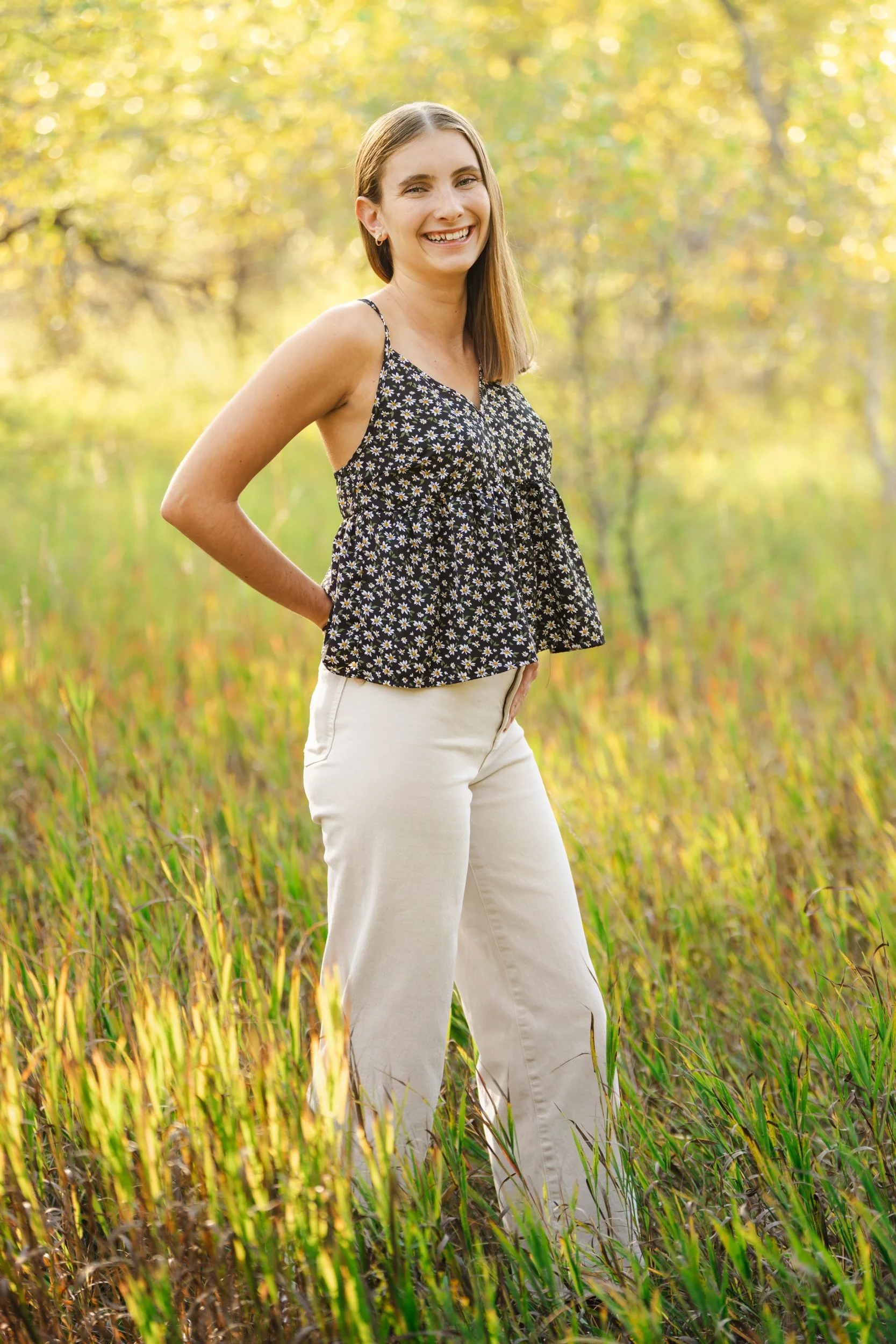 A young woman (high school senior) standing in a grassy field, smiling, wearing a black floral sleeveless top and white pants, with trees and sunlight in the background in Buffalo, Wyoming.