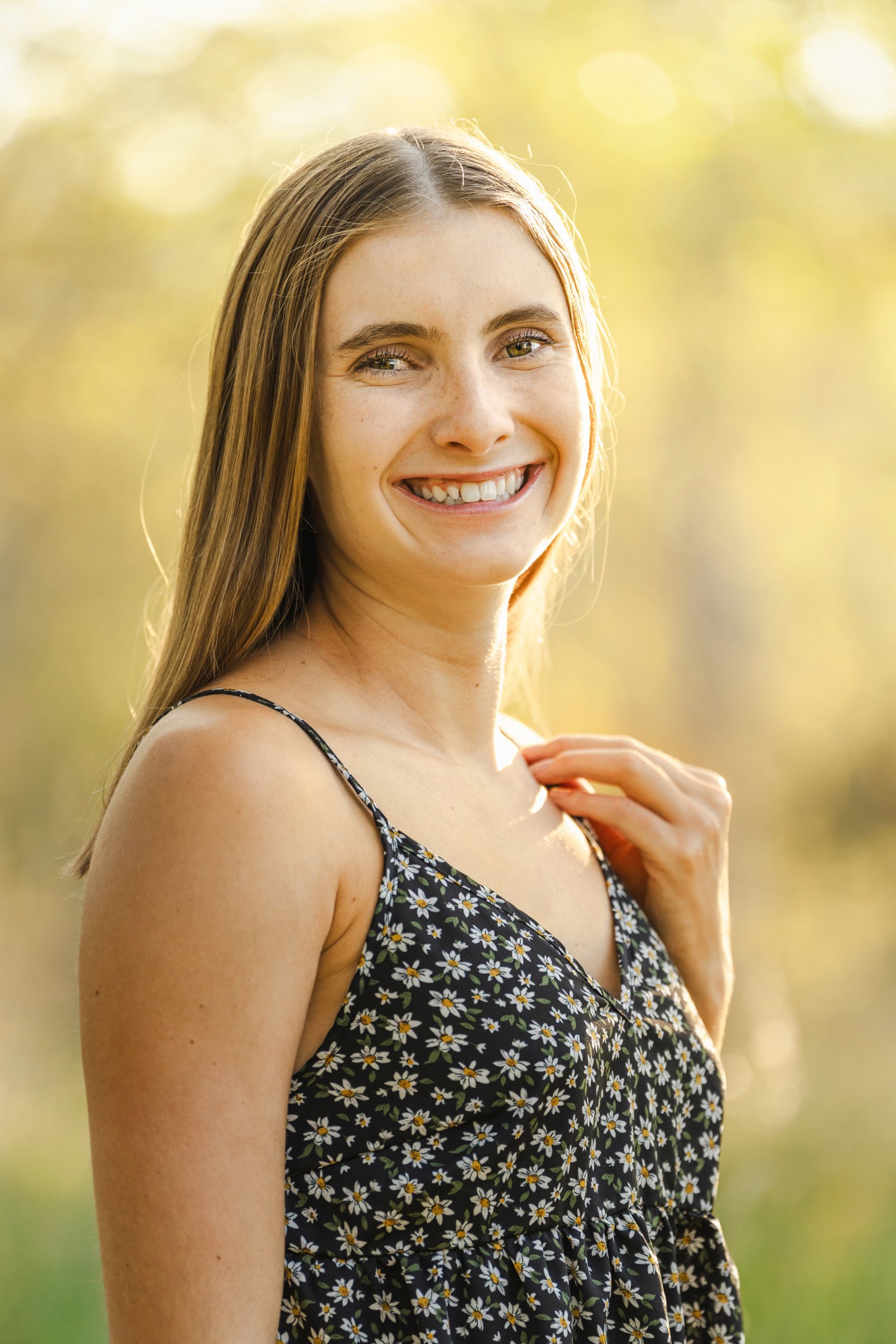 Smiling young woman (high school senior girl) with long light brown hair in a black tank top with white and yellow daisies standing outdoors in Buffalo, Wyoming with a blurred background of trees and sunlight.