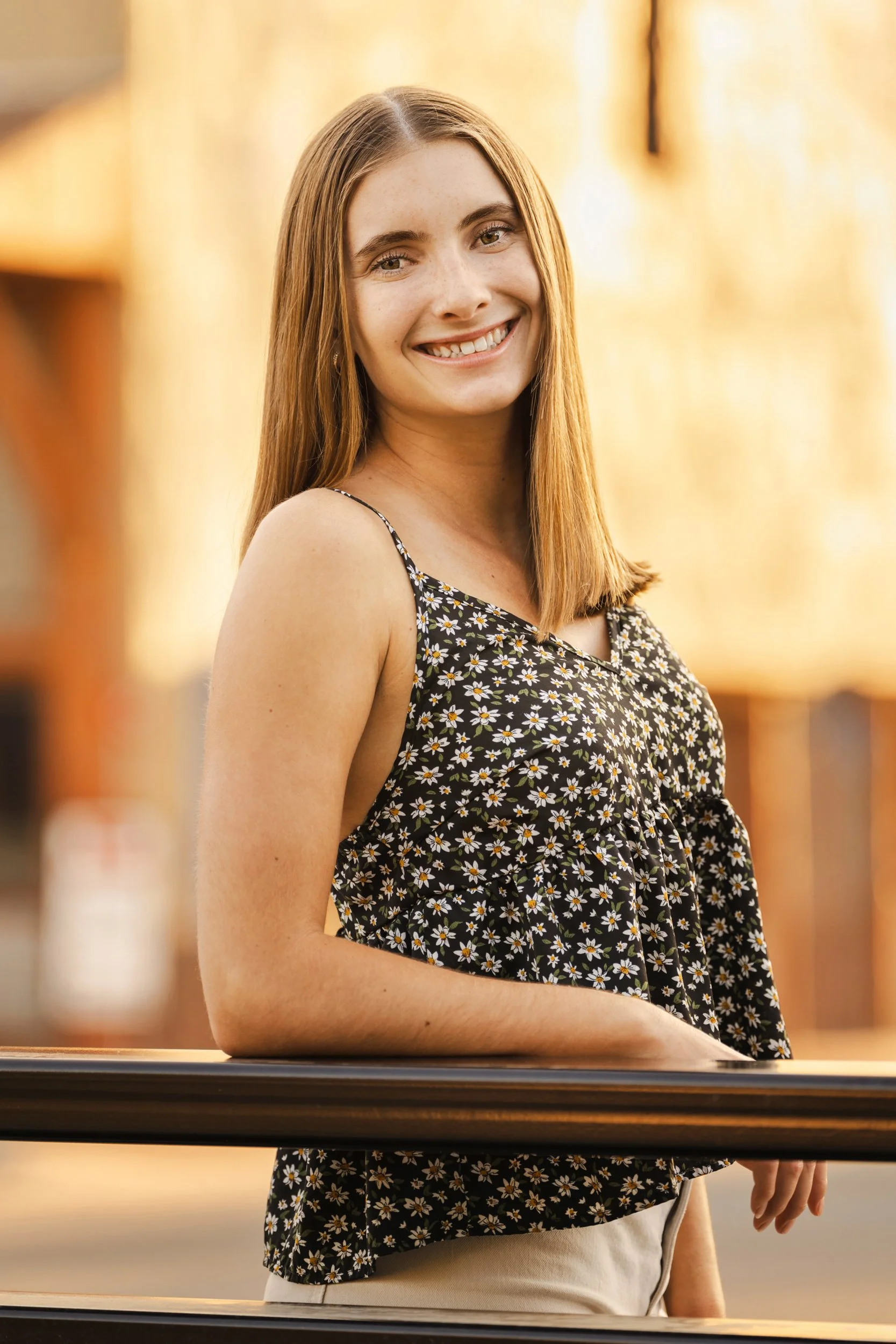 A Fort Collins, Colorado young woman (high school senior) with long hair smiling, wearing a black sleeveless floral top, standing outdoors in downtown Buffalo, Wyoming during golden hour.