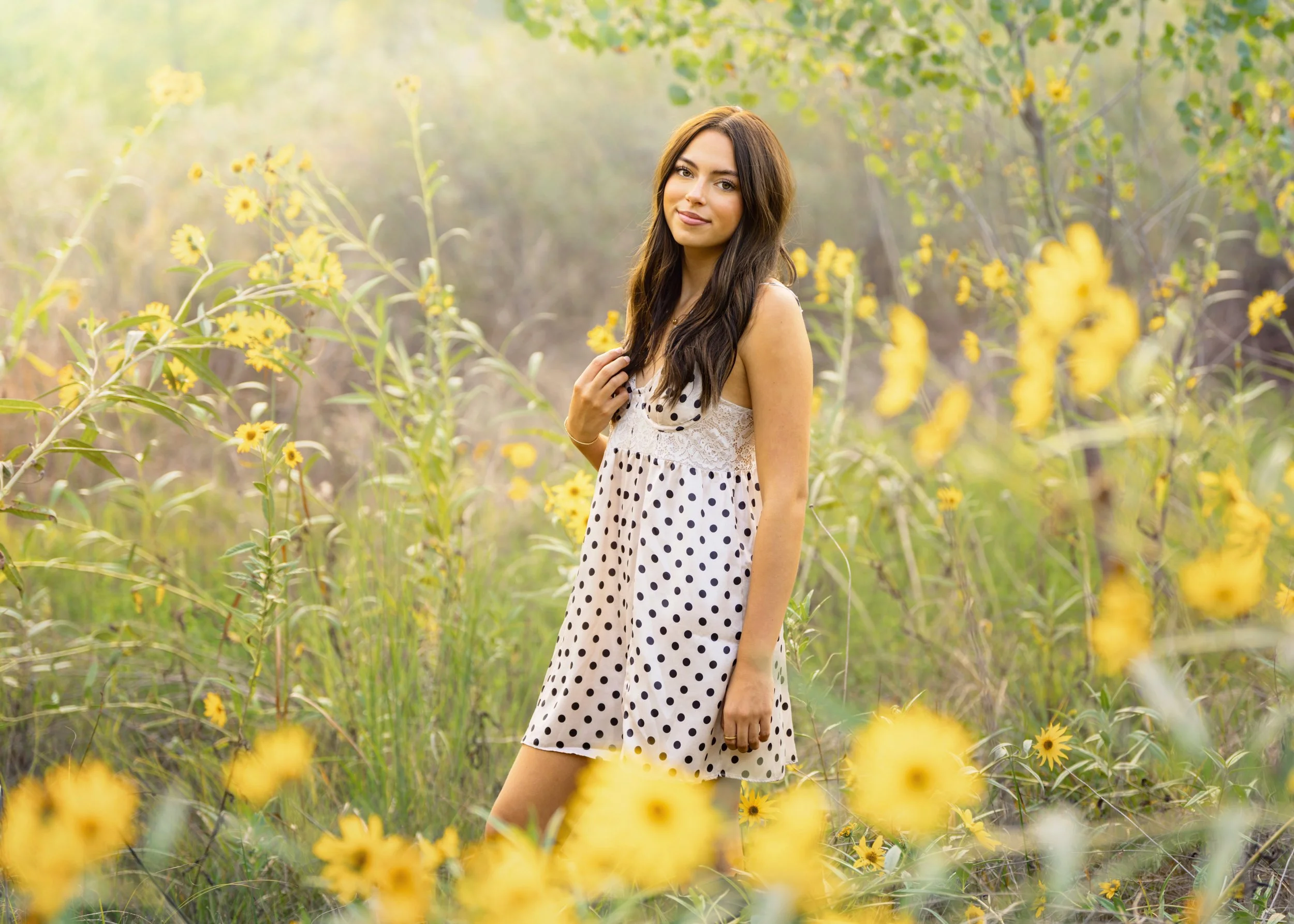 Young woman in a polka dot dress standing among yellow flowers in a field, with sunlight filtering through the trees in a Fort Collins, Colorado natural area for her high school senior photos.