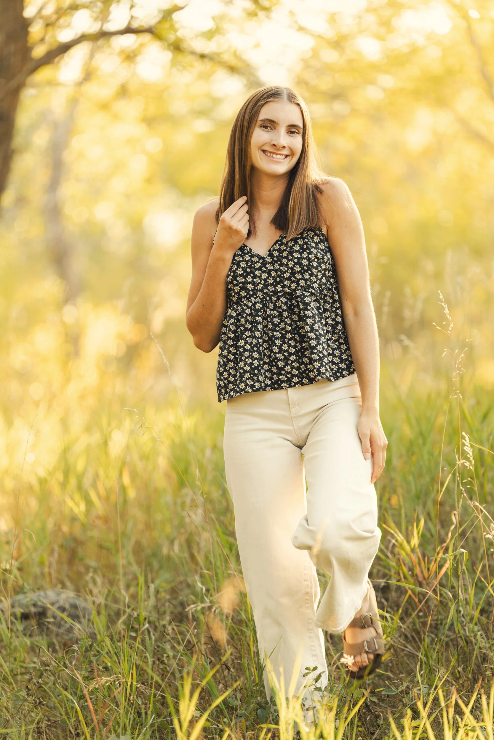 A high school senior girl with brown hair, wearing a black floral top, cream-colored pants, and sandals, smiling and walking through a grassy field in a forest during sunset in Buffalo, Wyoming.