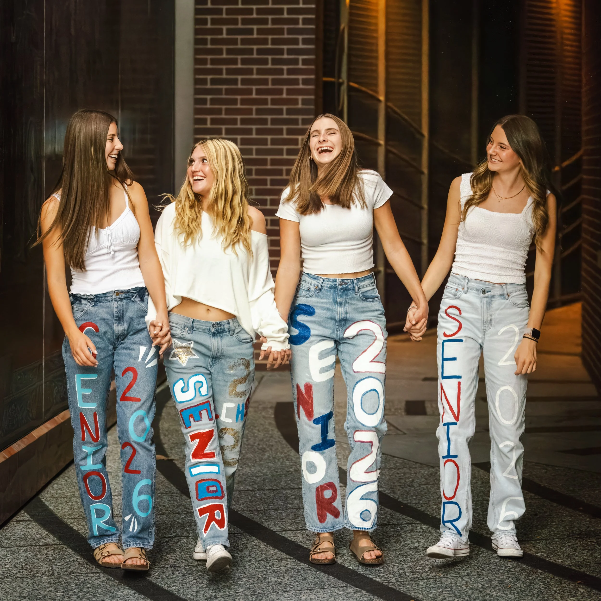 Four high school friends creating memories in white tops and decorated graduation jeans holding hands and walking outdoors in a downtown Ft. Collins, Colorado alley, smiling and enjoying themselves.
