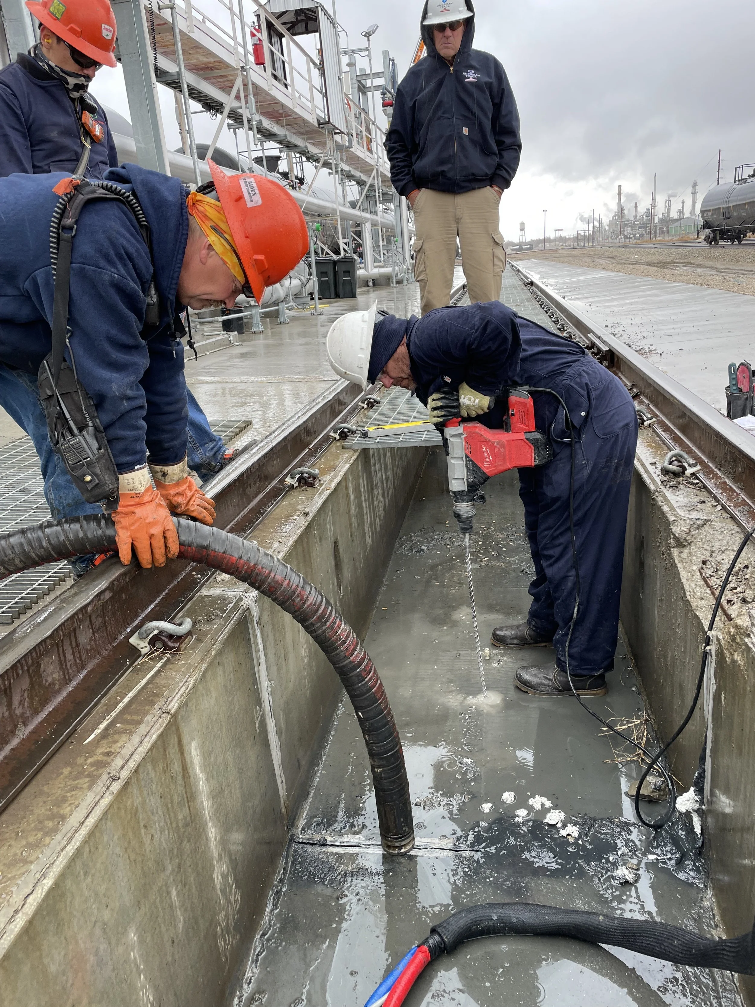 Workers in safety gear perform maintenance in a concrete trench next to railroad tracks at an industrial facility, with equipment and storage tanks visible in the background on a cloudy day.