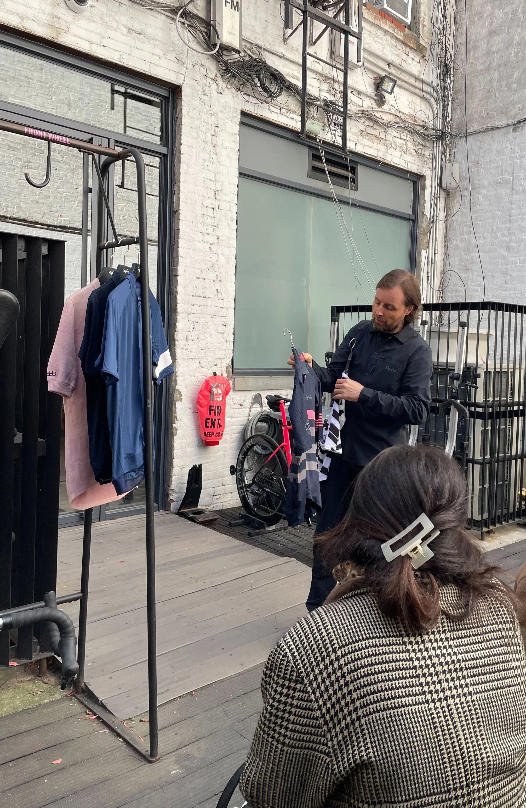Man holding clothes on a hanger showing to a woman seated outdoors at a clothing display outside a brick building.