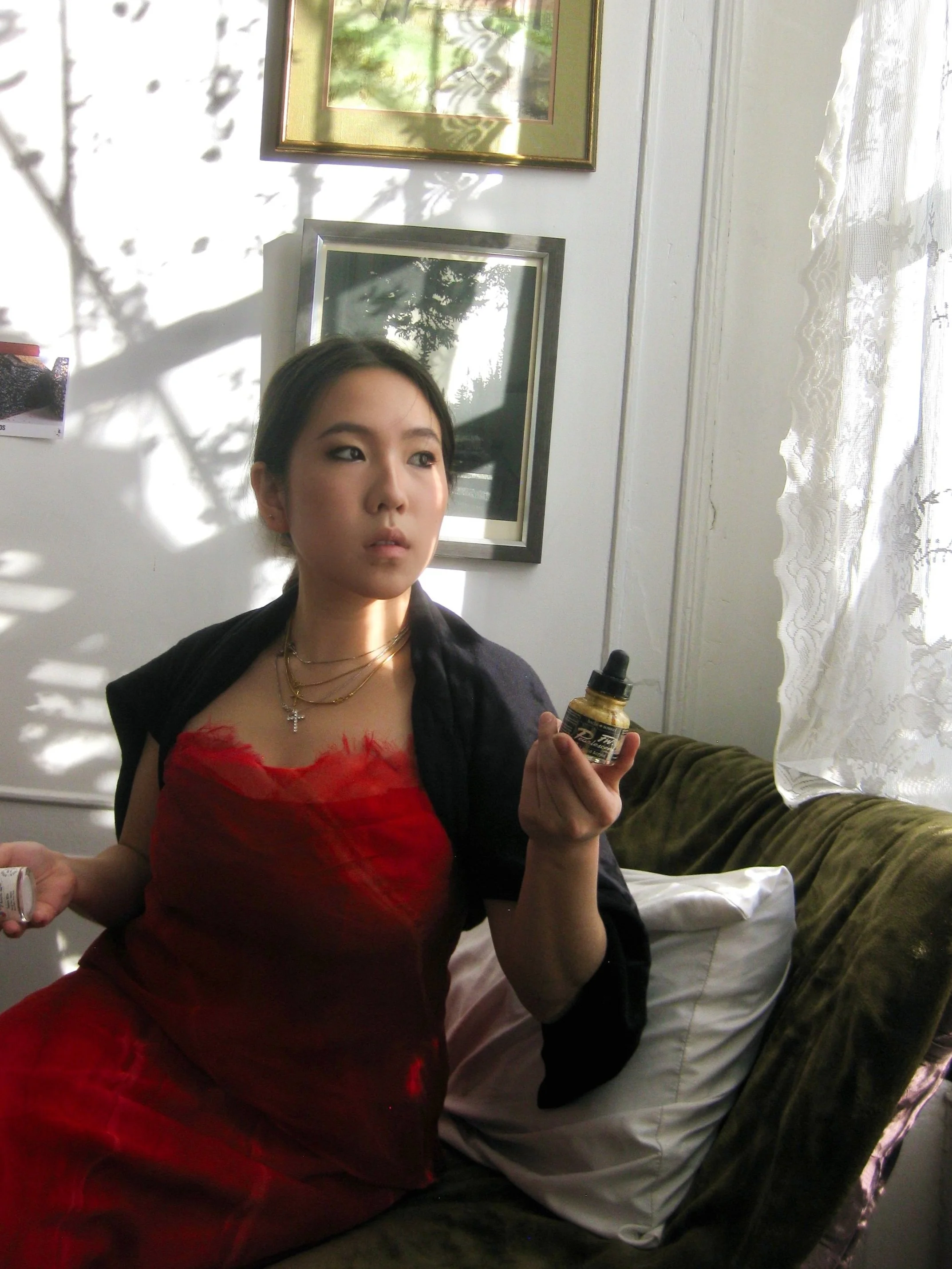 A woman with dark hair and dressed in a red top and a black jacket, sitting on a green couch in a well-lit room, holding a small bottle with a dropper in her right hand and another small bottle in her left hand. There are framed pictures on the wall behind her and a window with lace curtains to her right.