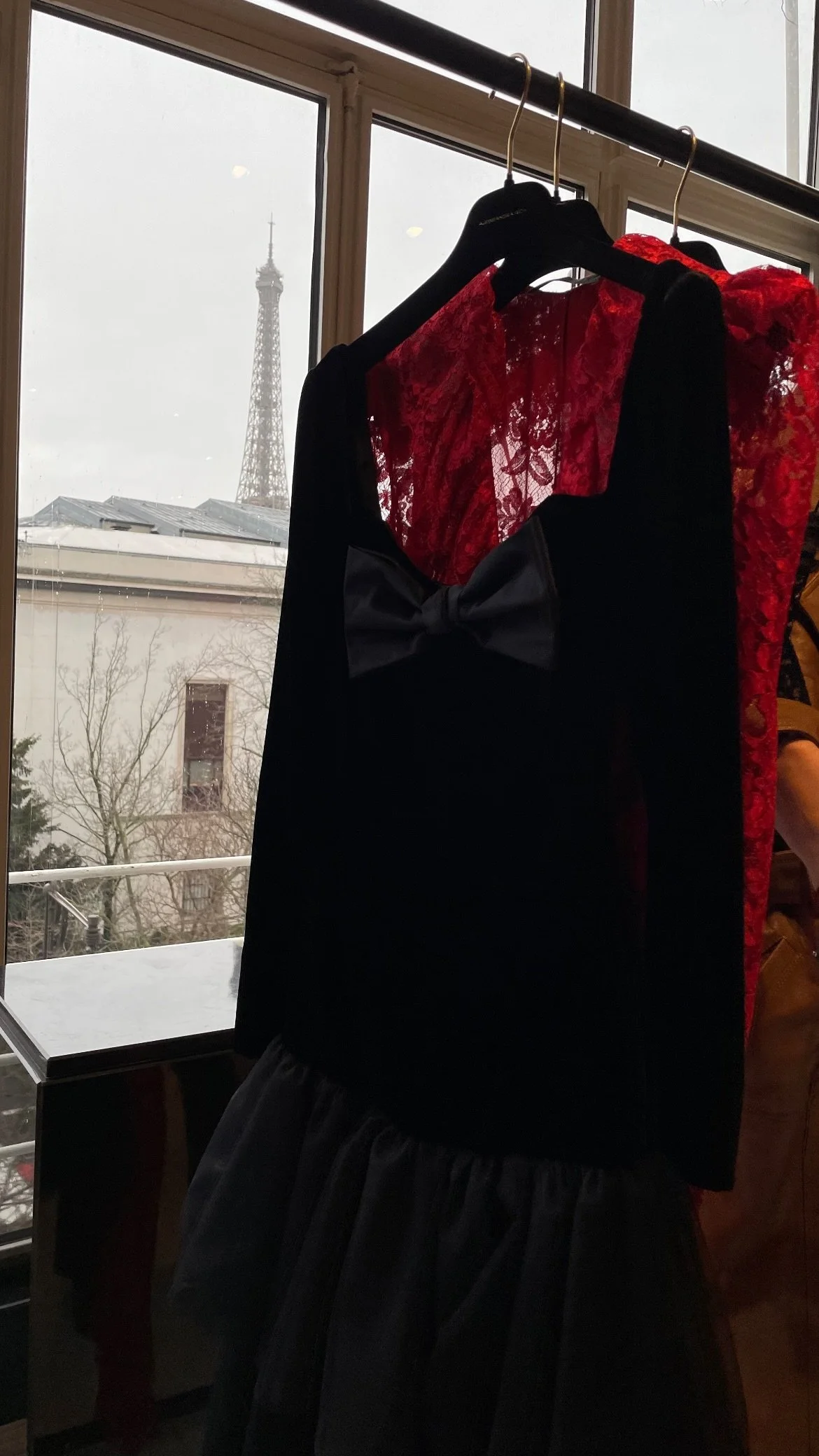 Fashion display with a black dress featuring a bow, a red lace dress, and a black and red outfit with a sheer red fabric, seen through a window with the Eiffel Tower in the background.