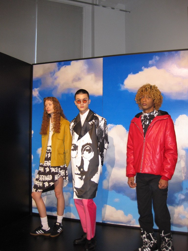 Three models posing in front of a painted backdrop of blue sky with white clouds, showcasing fashionable clothing and accessories.