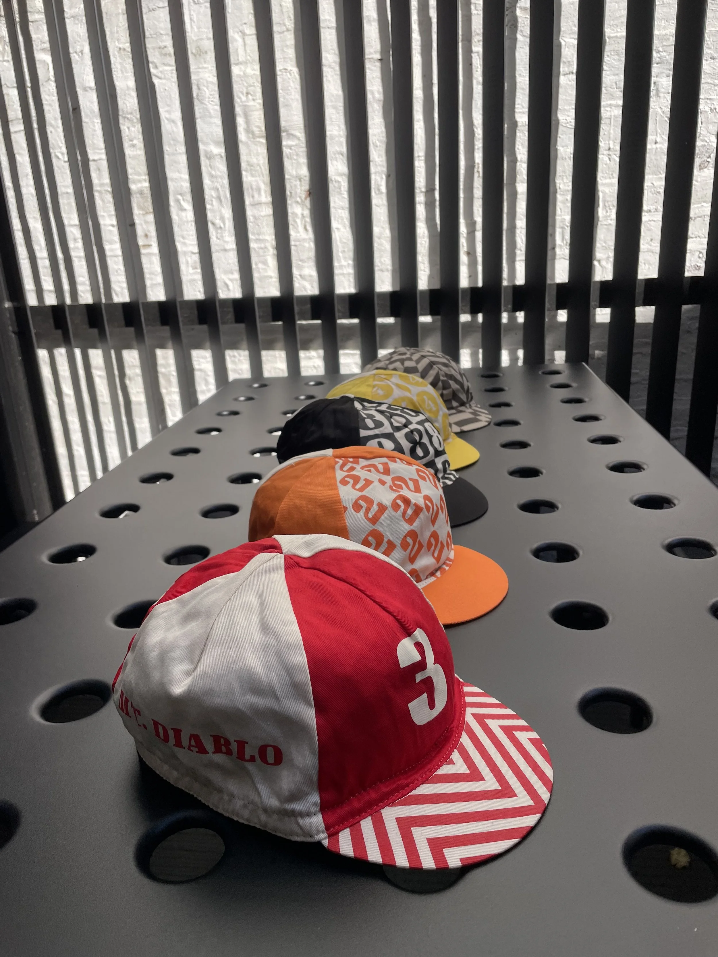 Five colorful baseball caps with various patterns, displayed on a gray perforated table against a background of black vertical bars and a white brick wall.