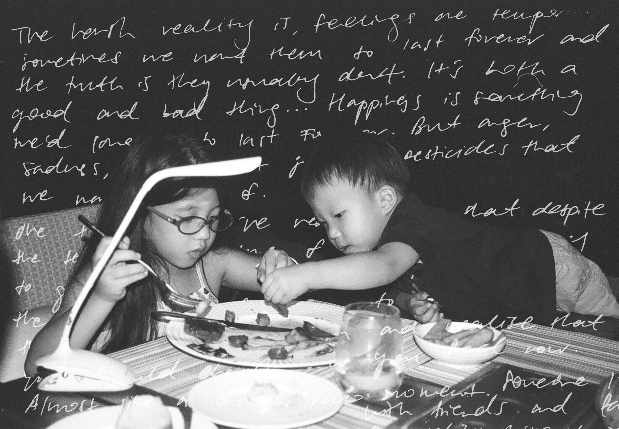 Black and white photo of two children, a girl with glasses and a boy, sharing food at a table with a lamp and writing on a blackboard in the background.