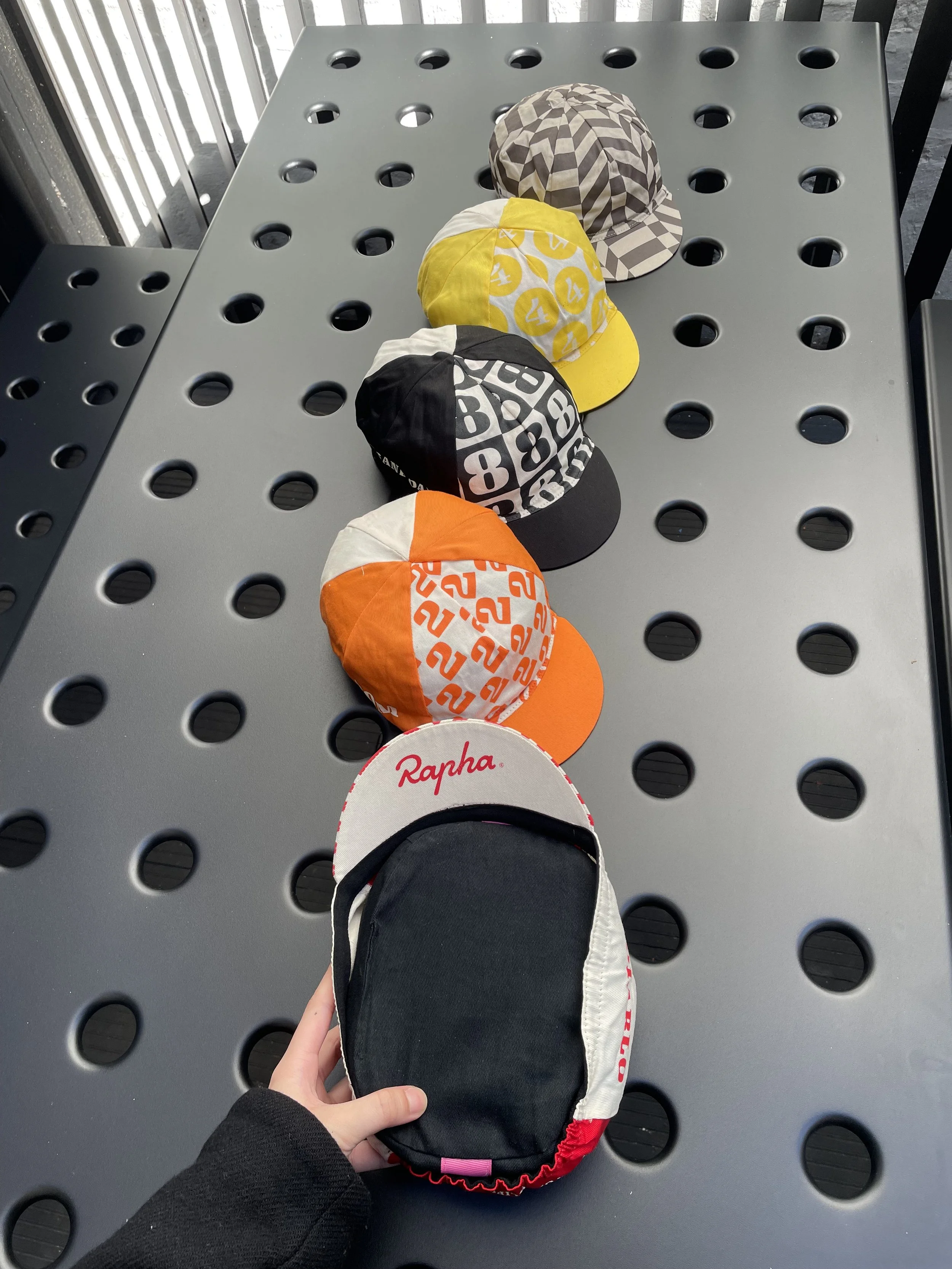 A row of five five-panel cycling caps with different colors and patterns on a perforated gray table, with a person holding a Rapha cycling cap in the foreground.